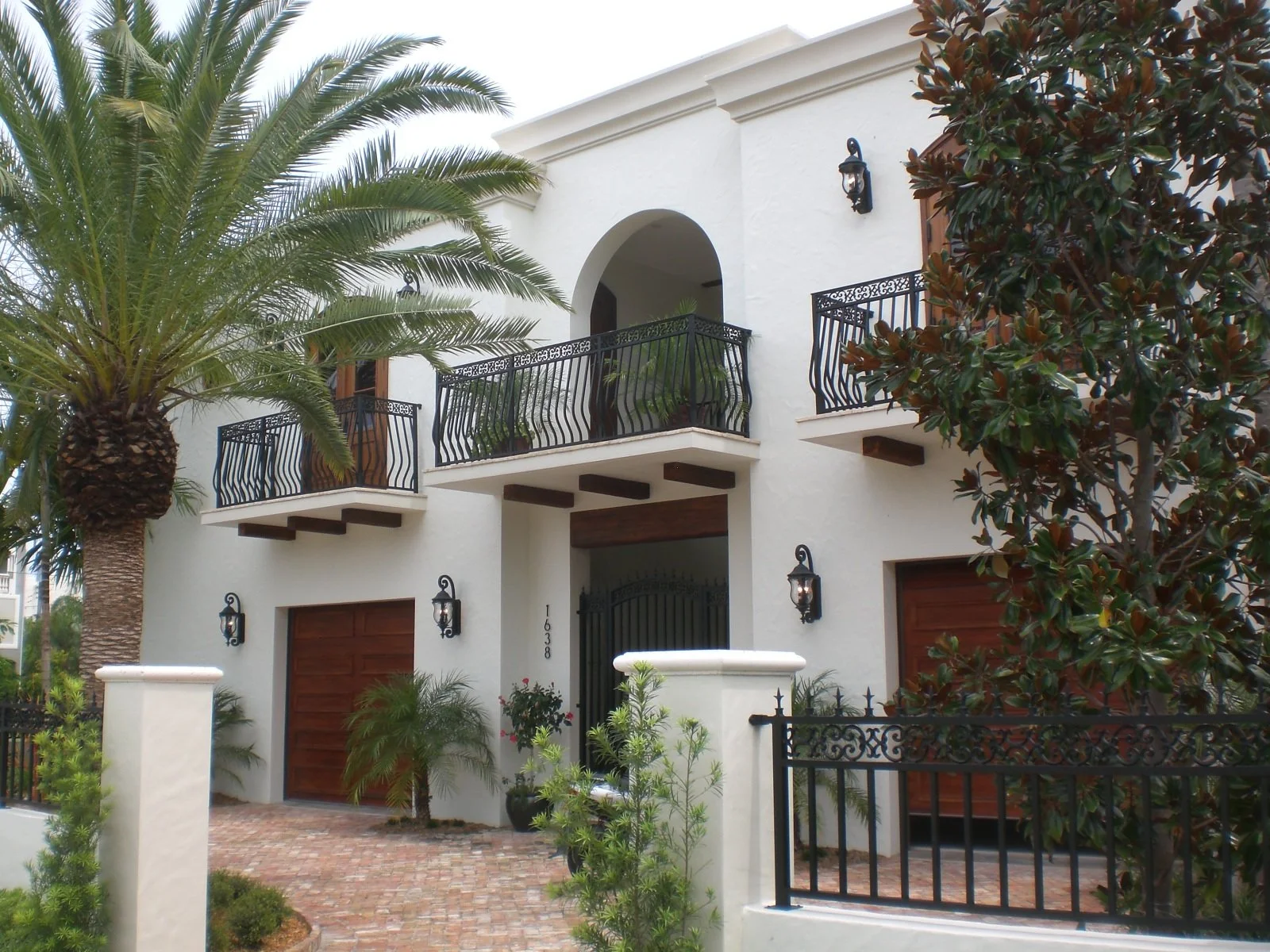 Front view of a two-story house with white stucco exterior, wooden garage doors, black wrought iron balcony and fence, tropical plants, palm tree, and wall-mounted lanterns.