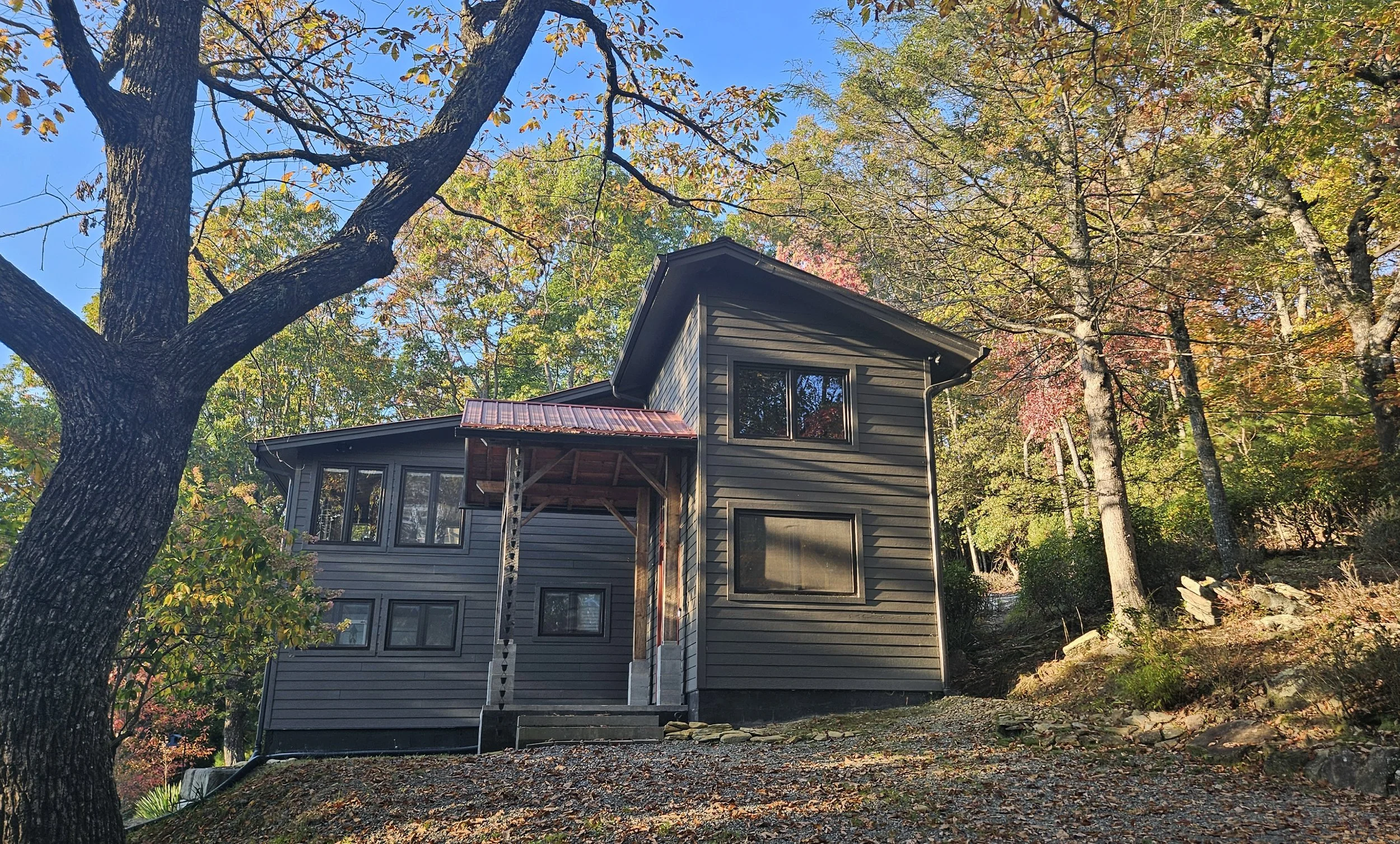 A two-story house with dark gray siding, large windows, and a metal roof, nestled among trees with autumn leaves.