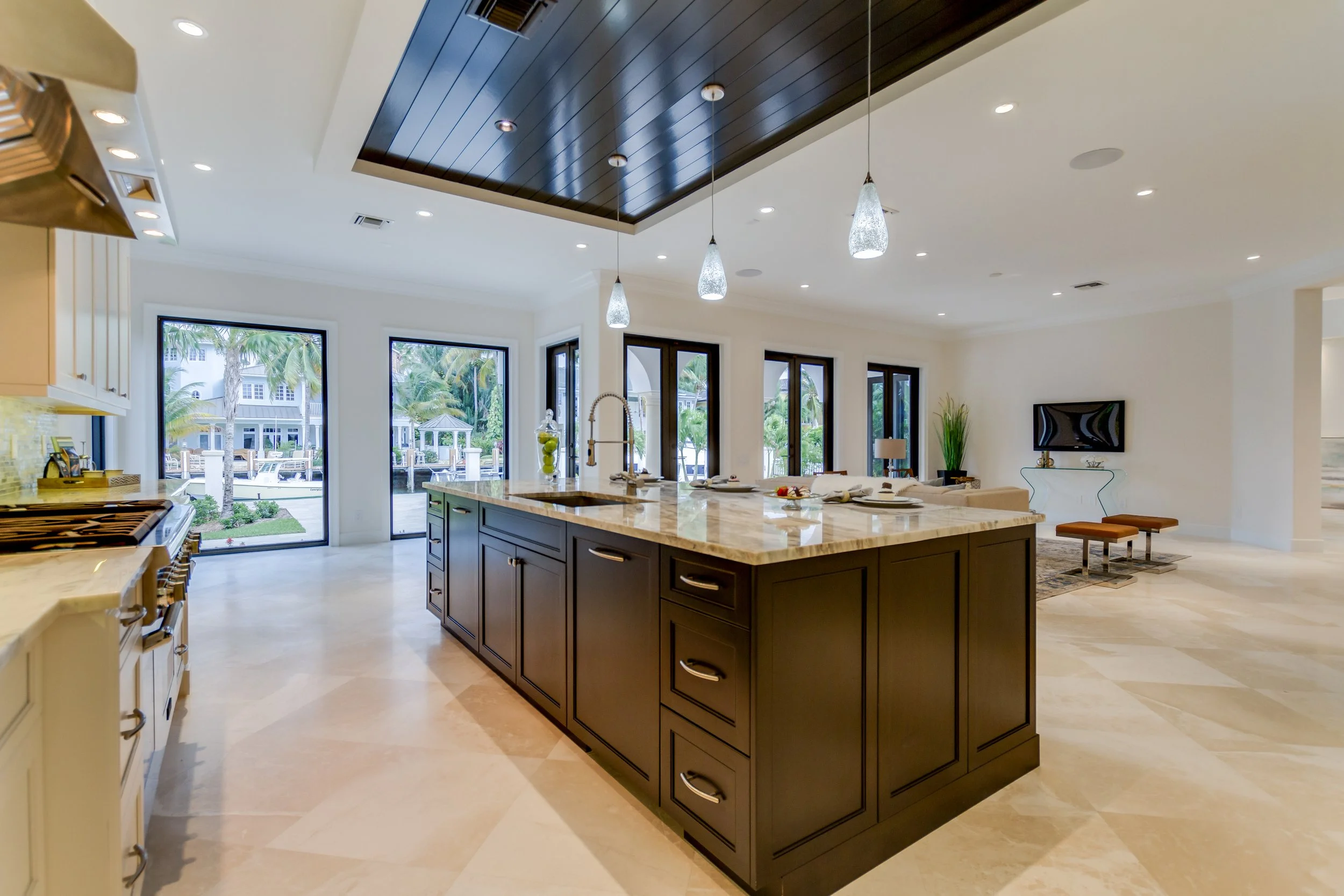Modern open-plan kitchen and living room with large windows, black kitchen island with marble countertop, beige tiled flooring, and a wall-mounted TV in the background.