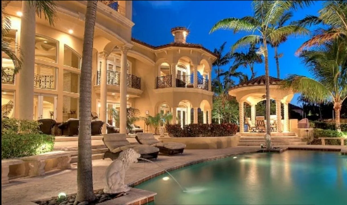 A luxurious mansion with a lit pool, surrounded by palm trees and outdoor seating areas at dusk.