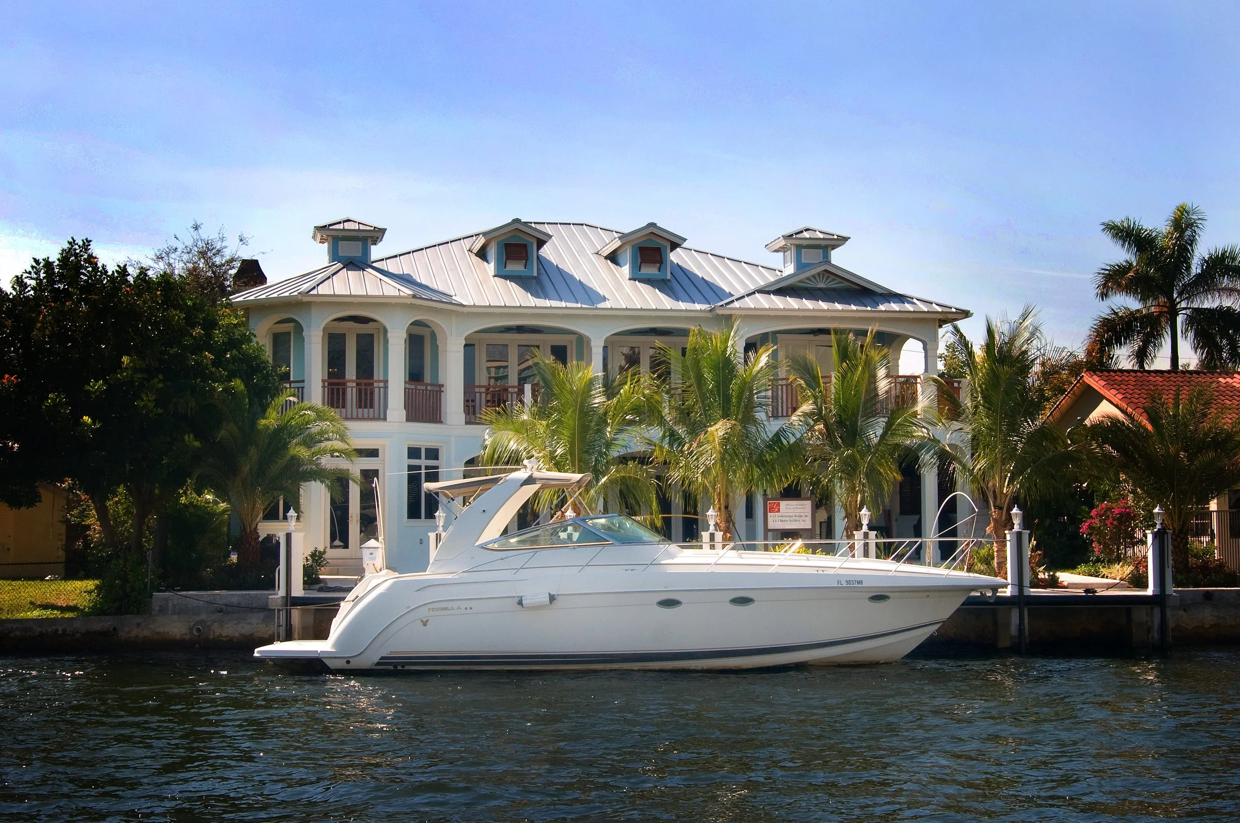 Luxury house with a white metal roof, large windows, and palm trees in front. A white yacht docked in the canal in front of the house.