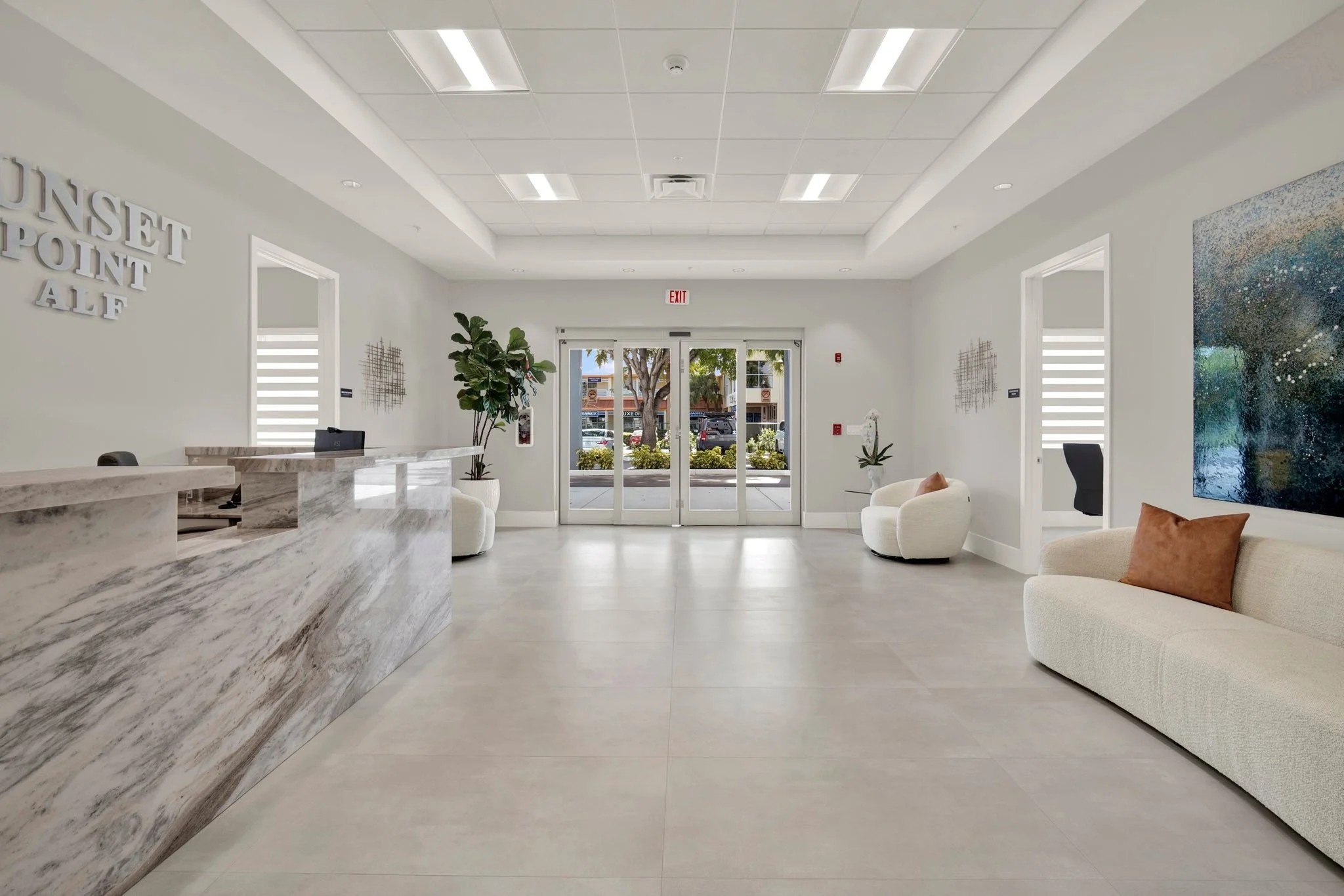 Modern, bright lobby with reception desk on the left, white walls, and seating area with sofas and armchairs. Large glass doors at the back open to outside view with trees and parked cars.
