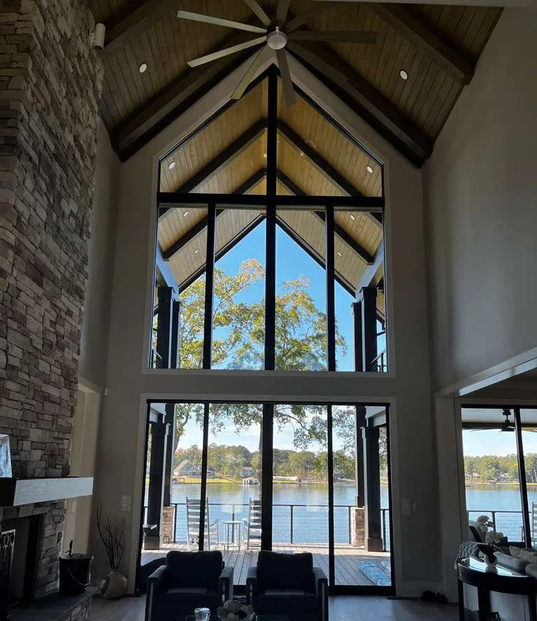 Interior view of a modern living room with a high vaulted ceiling, large floor-to-ceiling windows, and an outdoor patio overlooking a lake with trees.