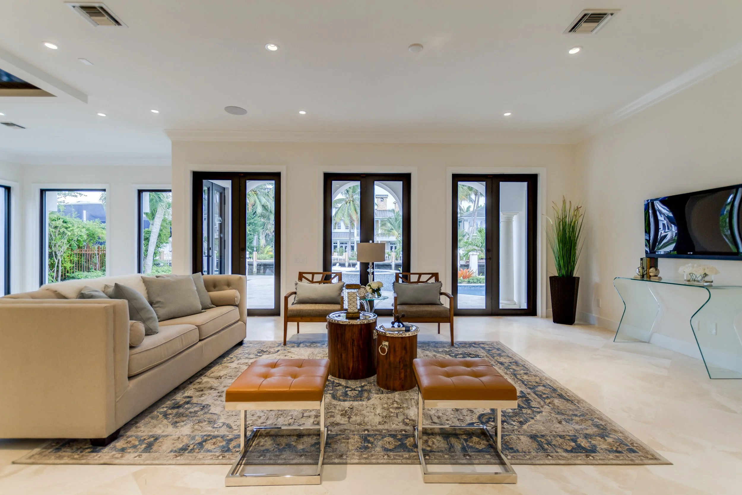 Modern living room with beige sofa, two wooden armchairs with cushions, two round wooden coffee tables with decorative items, two tan ottomans, a patterned area rug, large glass doors and windows overlooking tropical plants and palm trees, a tall pot