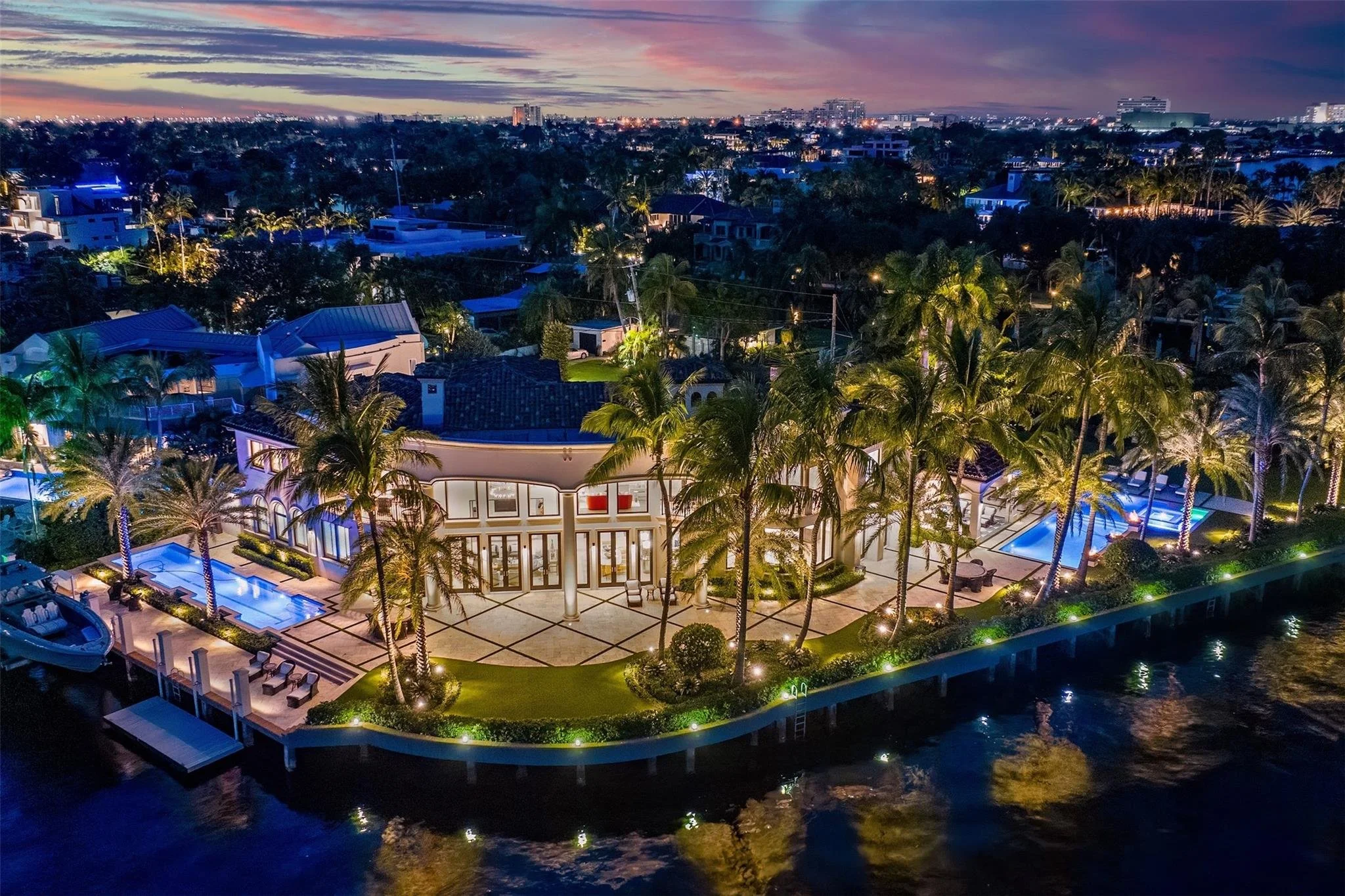 Aerial view of a luxurious waterfront property at sunset, featuring a modern house surrounded by palm trees, a swimming pool, and outdoor seating areas, with city lights visible in the distance.