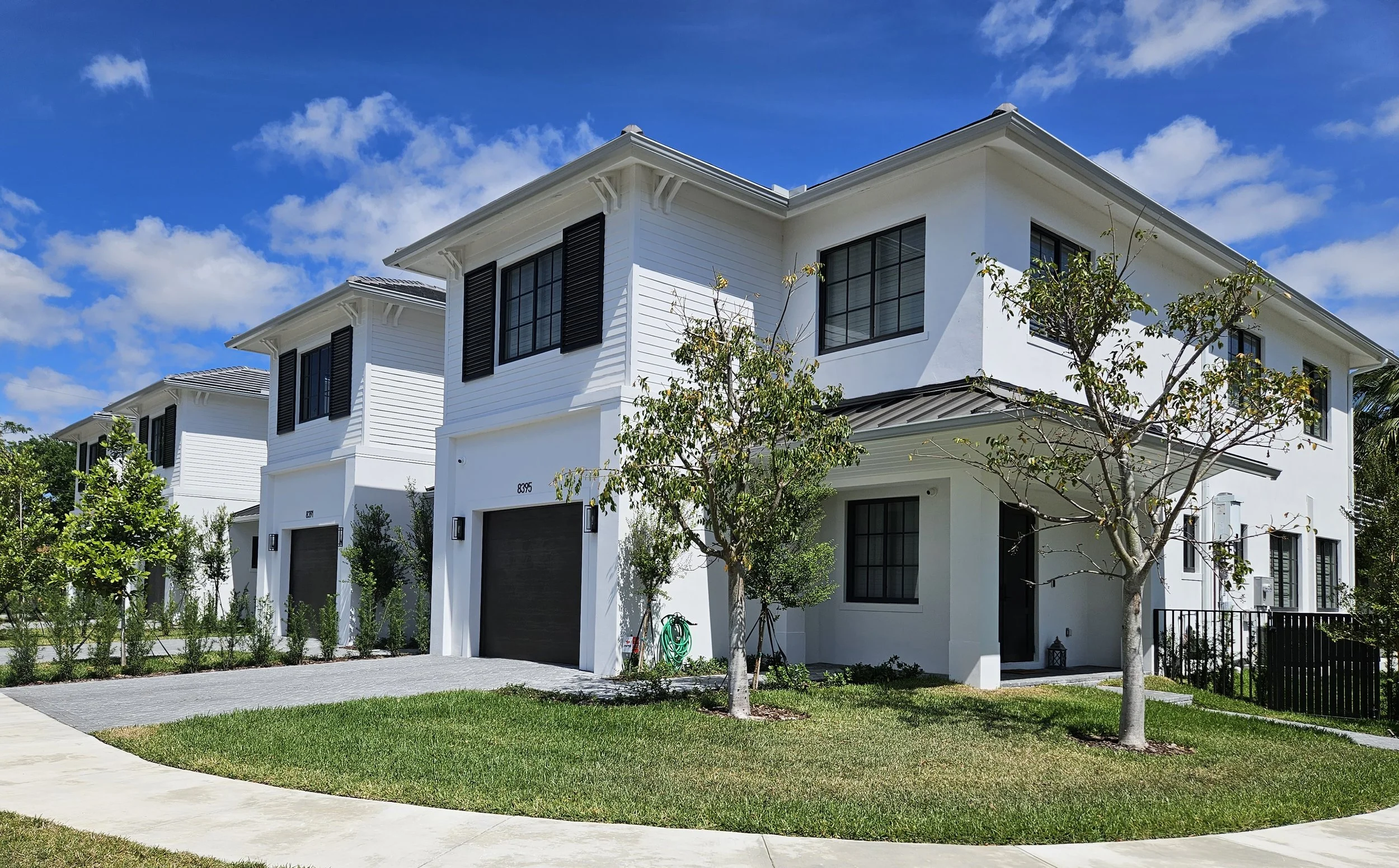 Modern white residential townhouses with black shutters, front yard with grassy lawn, young trees, and a concrete sidewalk under a partly cloudy blue sky.