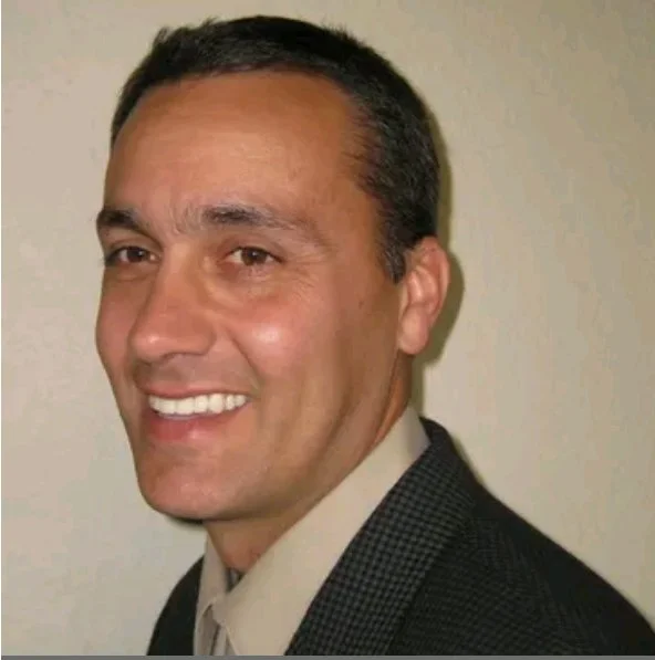 A man in a suit smiling, with short dark hair and a light complexion, standing against a plain background.