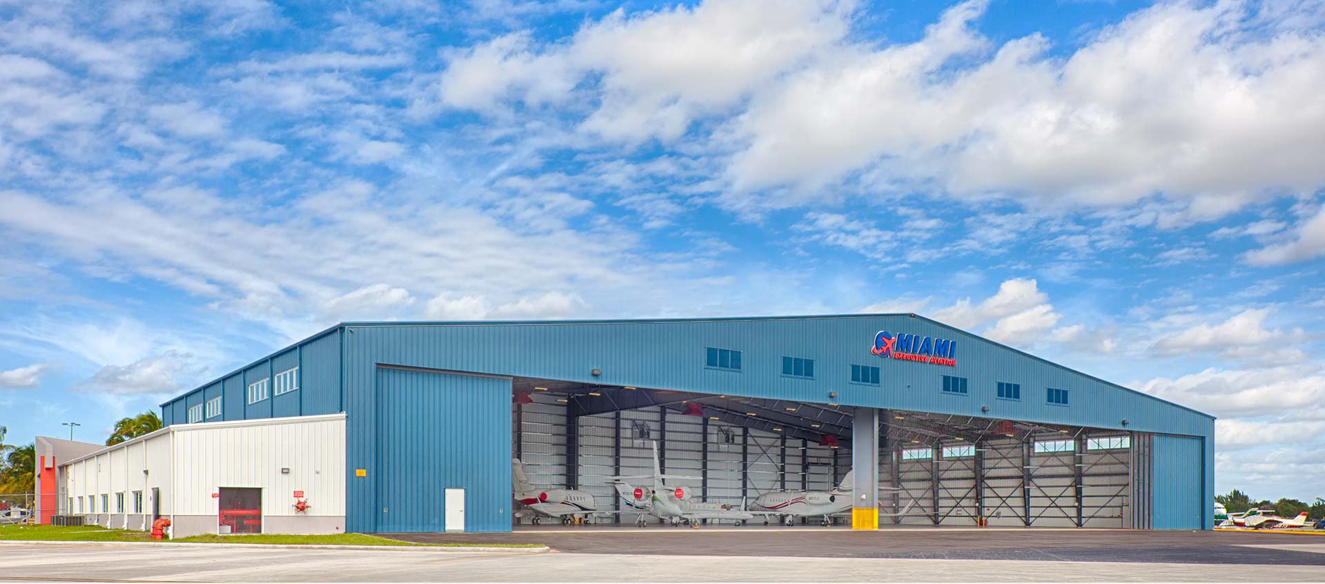Large aircraft hangar with an open front showing several small private jets parked inside, blue exterior with a Miami Aviation sign, under a partly cloudy sky.