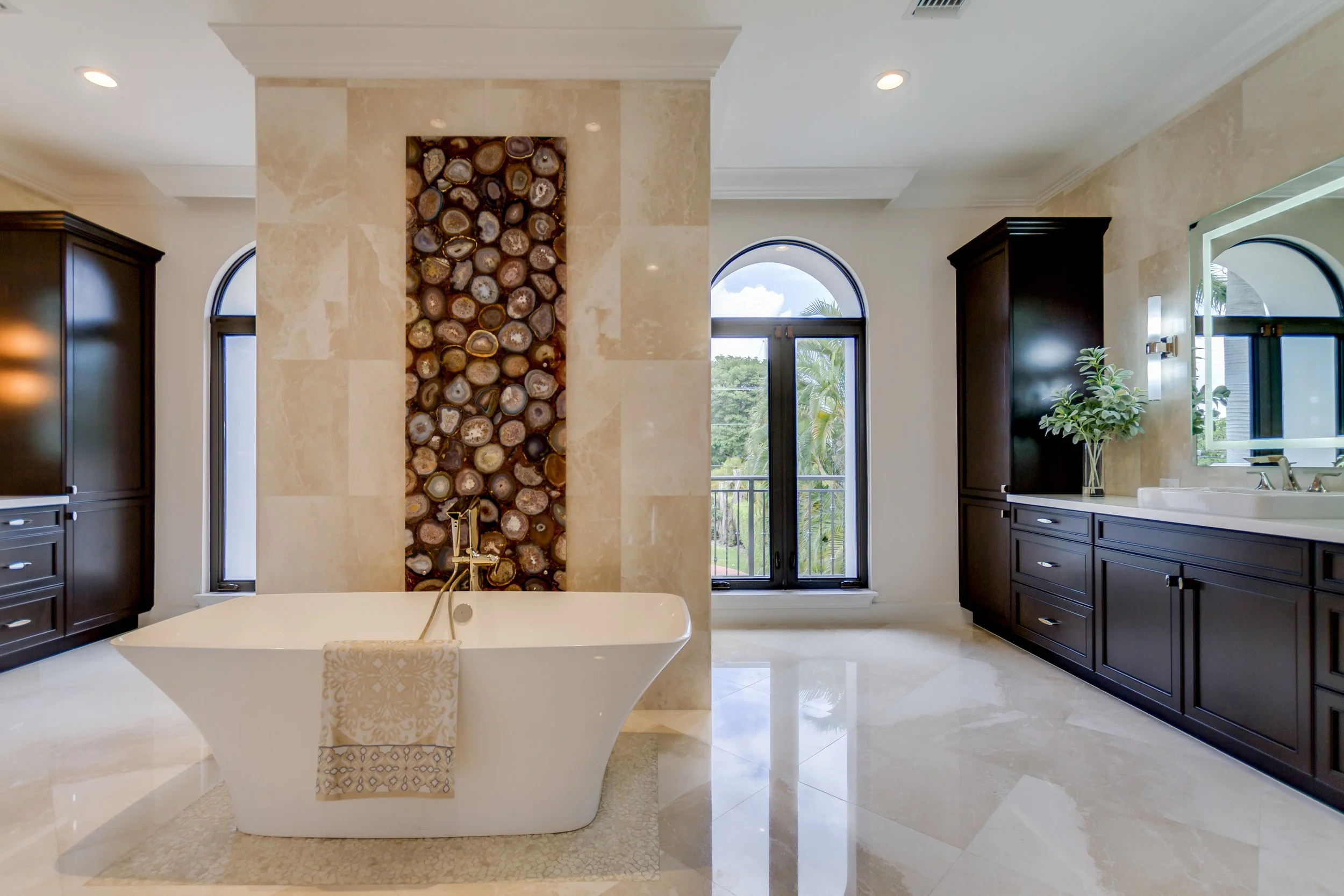 A modern bathroom with a white freestanding bathtub in front of a decorative wall with stacked logs. The room has beige marble flooring, dark wooden cabinetry, a large mirror, and arched windows with black frames showing greenery outside.
