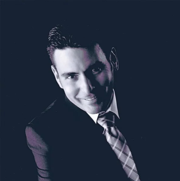 A man smiling in a professional headshot, wearing a dark suit and a plaid tie, against a dark background.