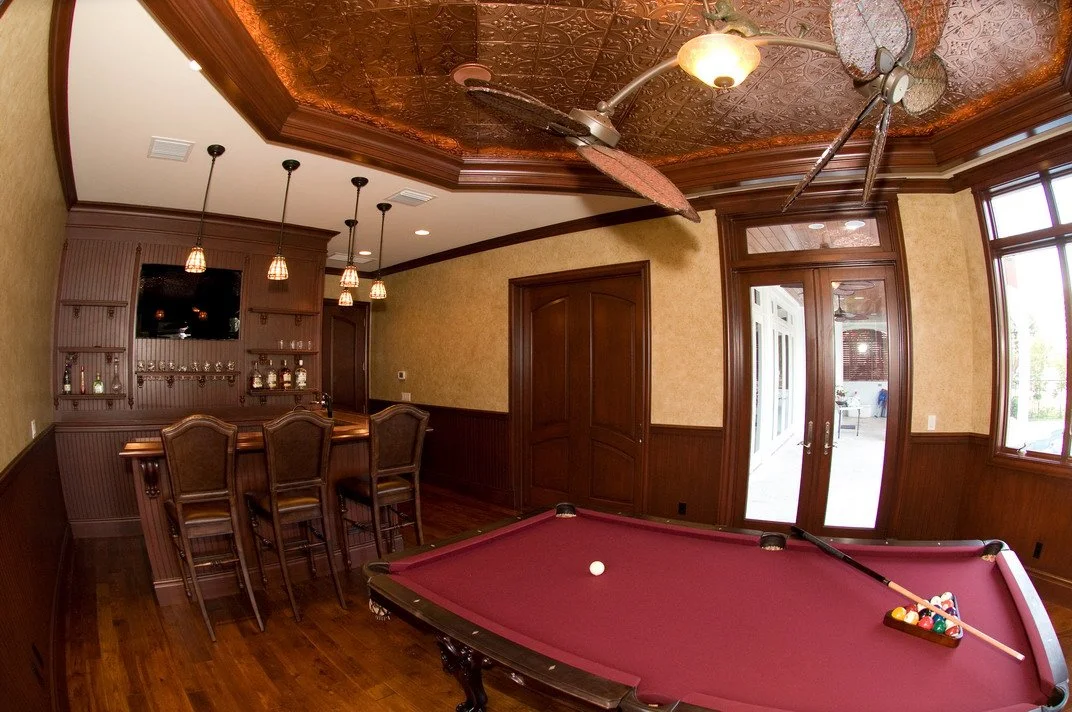 A billiards table with a ball and a cue stick, a wet bar with chairs, and a ceiling fan in a home game room with wood paneling and windows.
