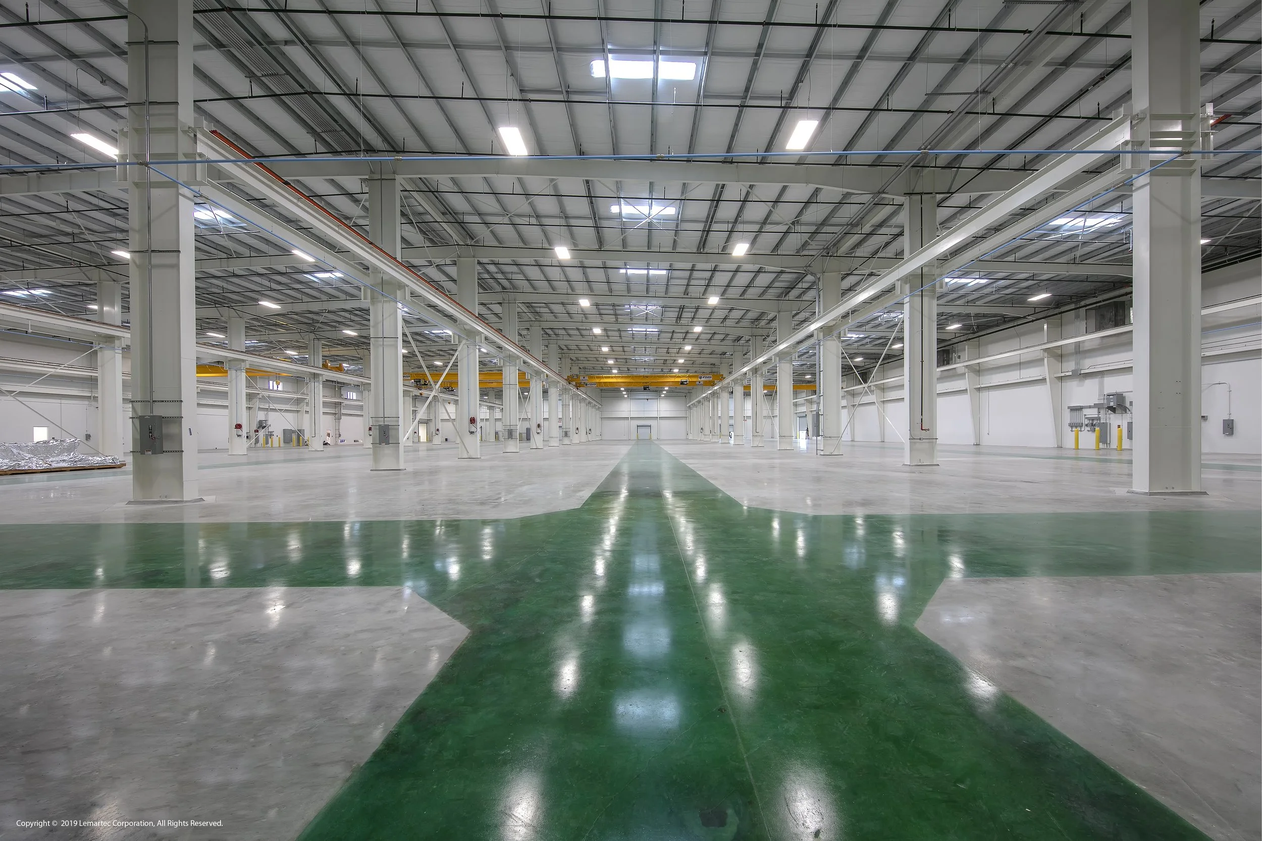 Empty warehouse with a green arrow painted on the floor pointing straight ahead, high ceilings, metal support beams, and overhead lighting.
