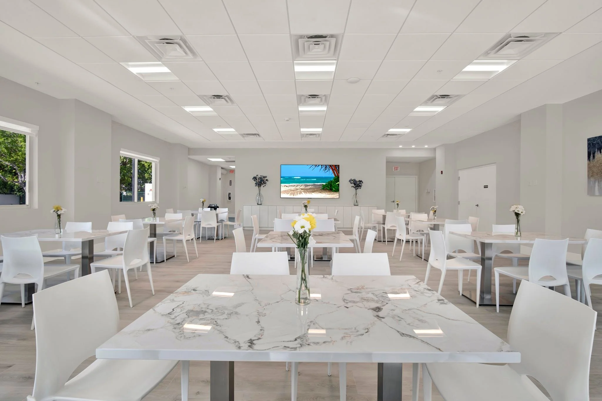 A bright, modern dining area with white tables, chairs, and a large wall-mounted TV displaying a beach scene. Flowers in vases decorate each table, and windows let in natural light.