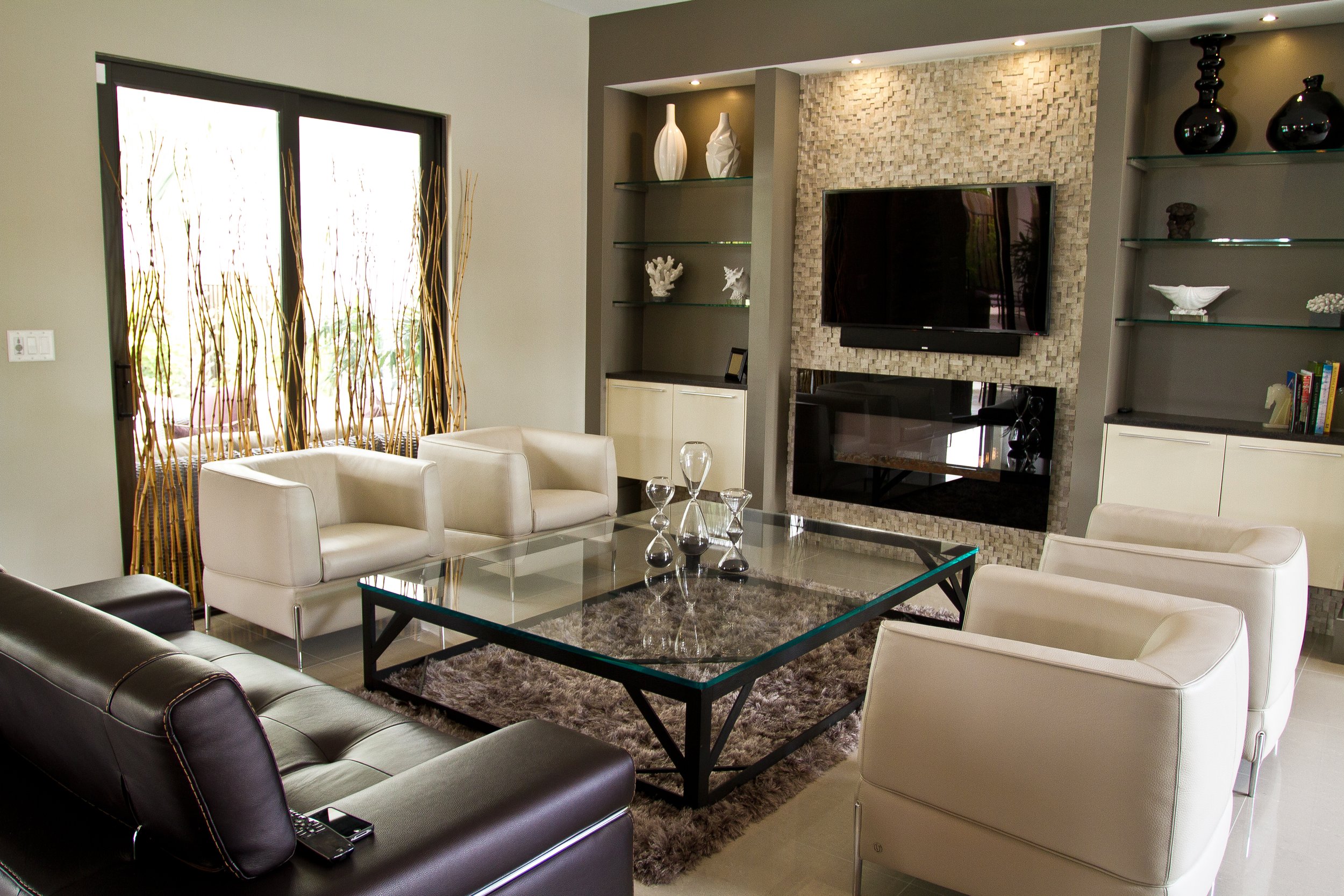Modern living room with a glass coffee table, white and beige armchairs, a black leather sofa, a textured stone fireplace with a television, and built-in shelves with decorative items.