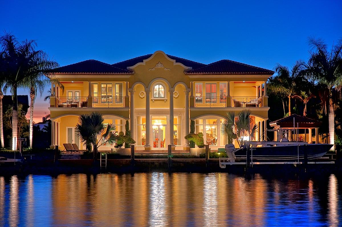 Luxury waterfront house illuminated at dusk with palm trees, a boat docked nearby, and calm water reflecting the house and sky.