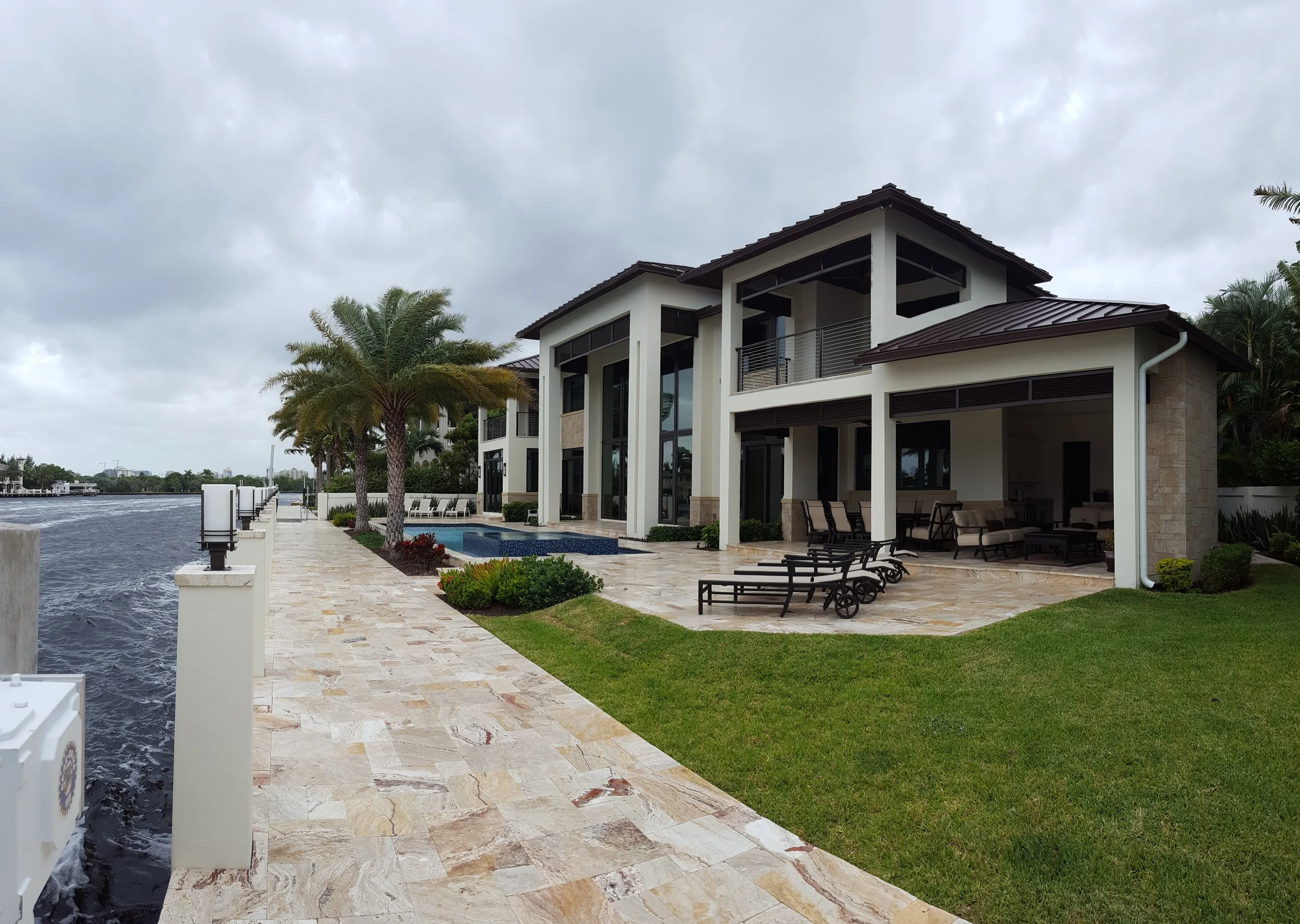 Modern waterfront house with a patio, lounge chairs, palm trees, and a canal, on a cloudy day.