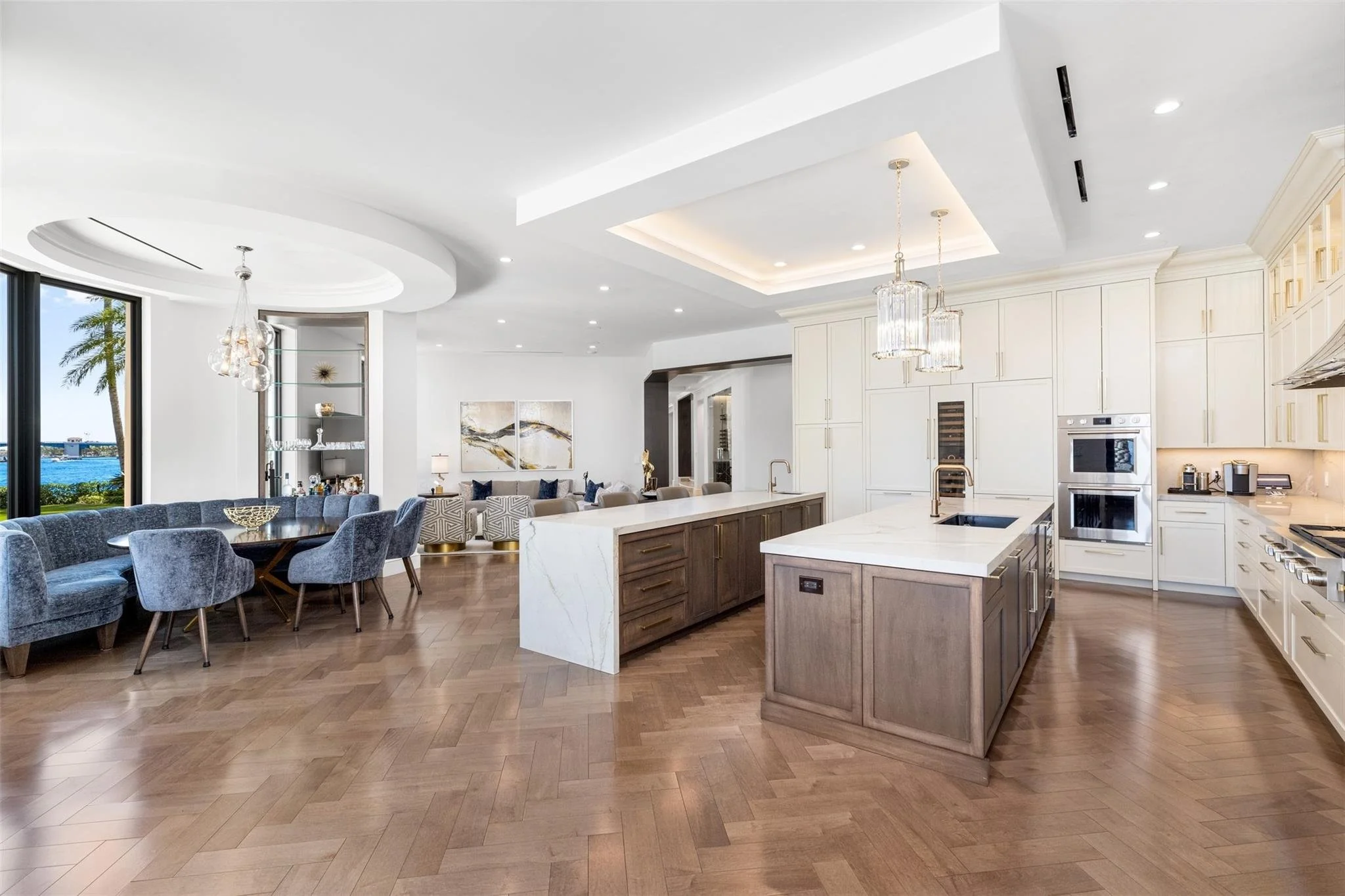 Open-concept kitchen and living area with white cabinetry, wooden flooring, and large windows showing outdoor scenery with palm trees and water.