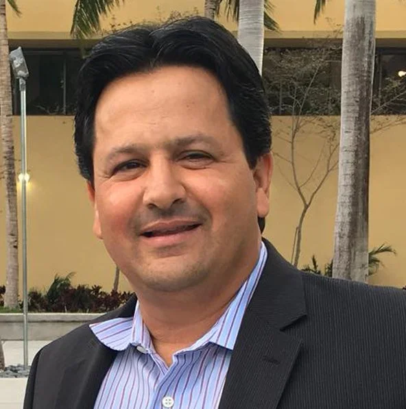 A man with dark hair, dressed in a striped shirt and dark blazer, standing outdoors with palm trees and a building in the background.