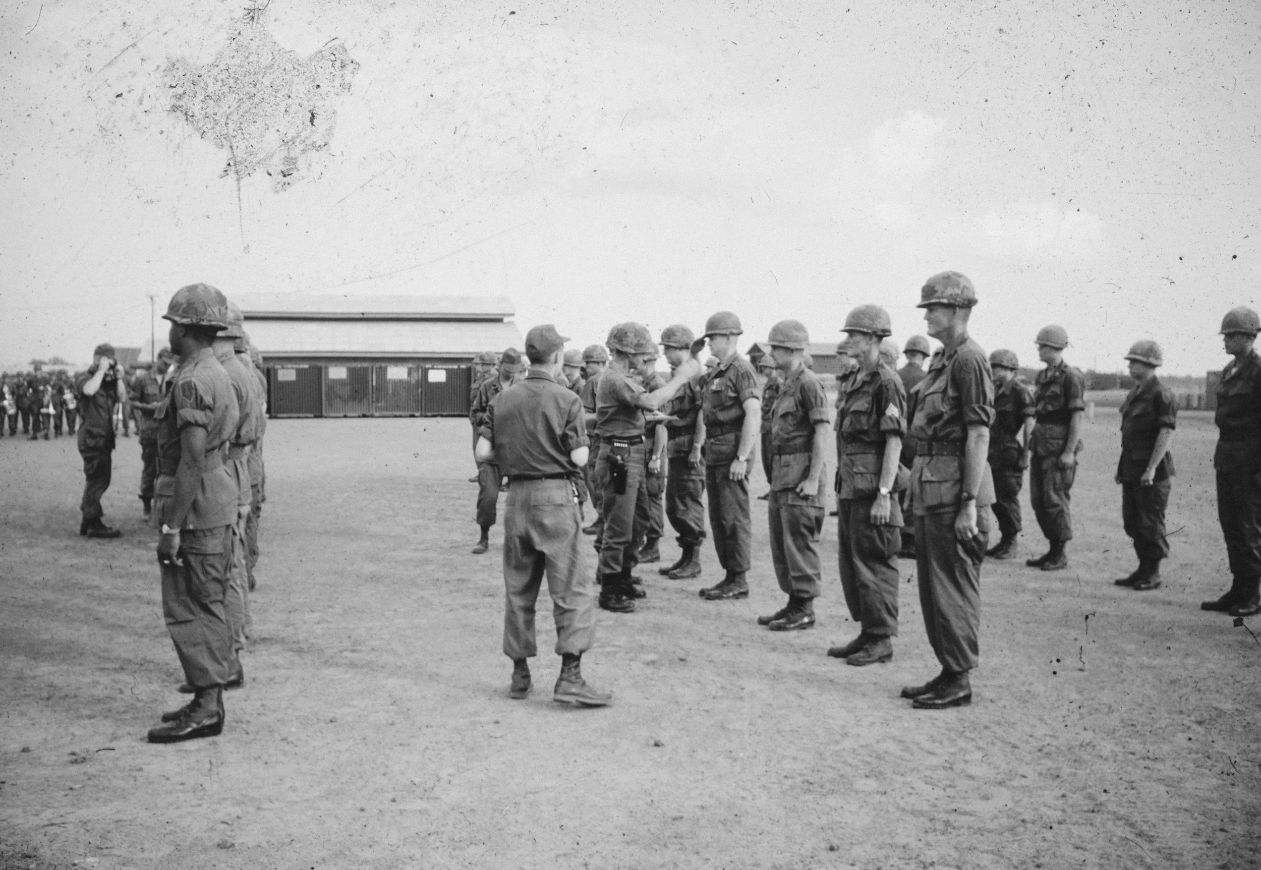 Black and white photo of soldiers in uniform with helmets, standing in formation outdoors, with a person in uniform presenting a medal or award.