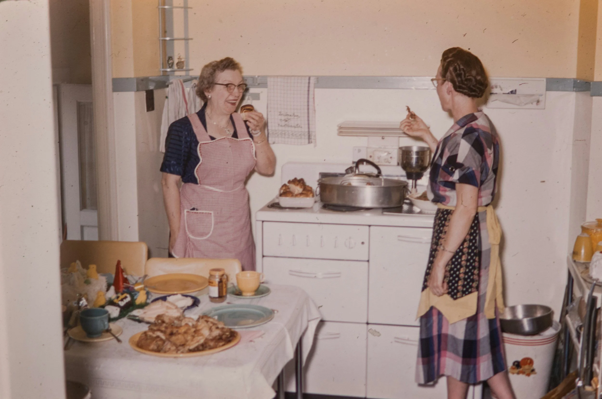 Two women with aprons in a kitchen, one is holding a fork and smiling, the other is eating a slice of pie, a table with food and dishes is in front of them.