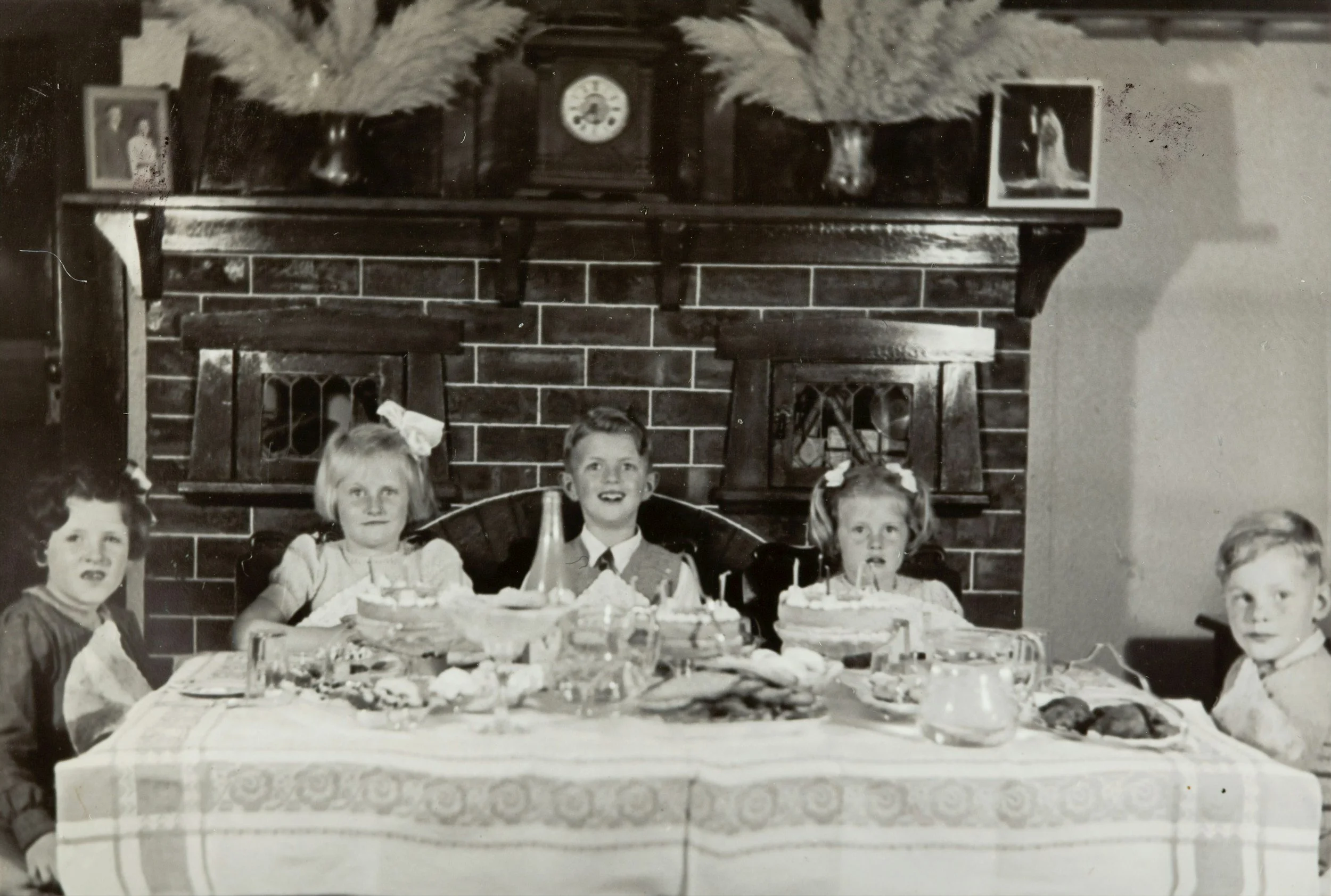A vintage black and white photo of five children sitting at a birthday party table with cakes, snacks, and drinks in front of a brick fireplace with a clock and decorative items.