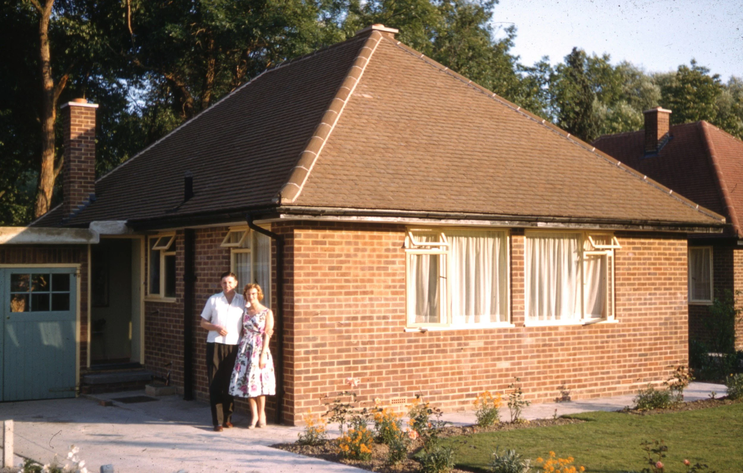A brick house with a sharply sloped roof, three open windows with curtains, and two people standing in front of the house, outdoors on a sunny day.