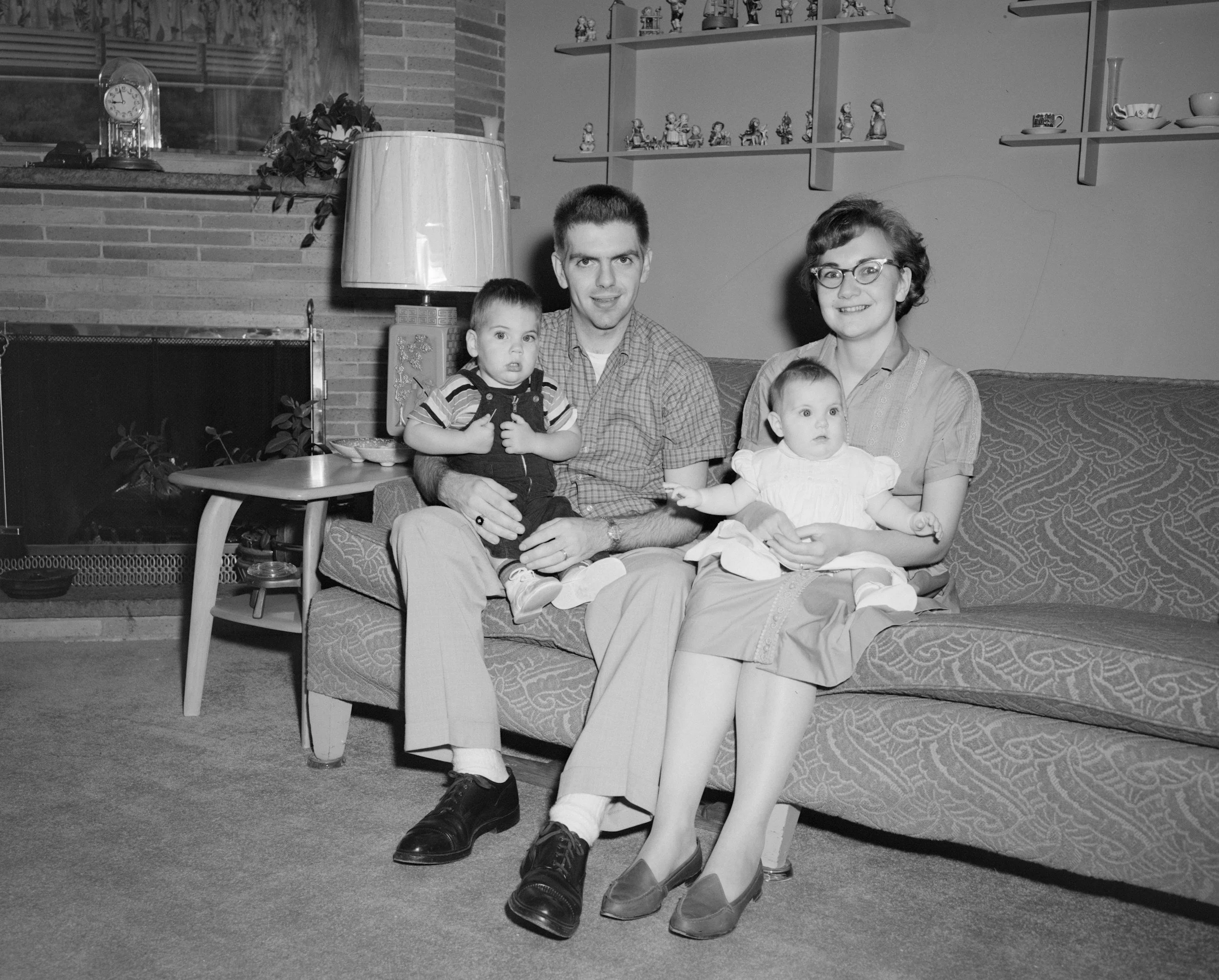 A black and white photo of a family sitting on a patterned sofa in a living room. The man is holding a young boy, and a woman is holding a baby girl. There are shelves with small figurines, a lamp, and a clock on a brick fireplace in the background.