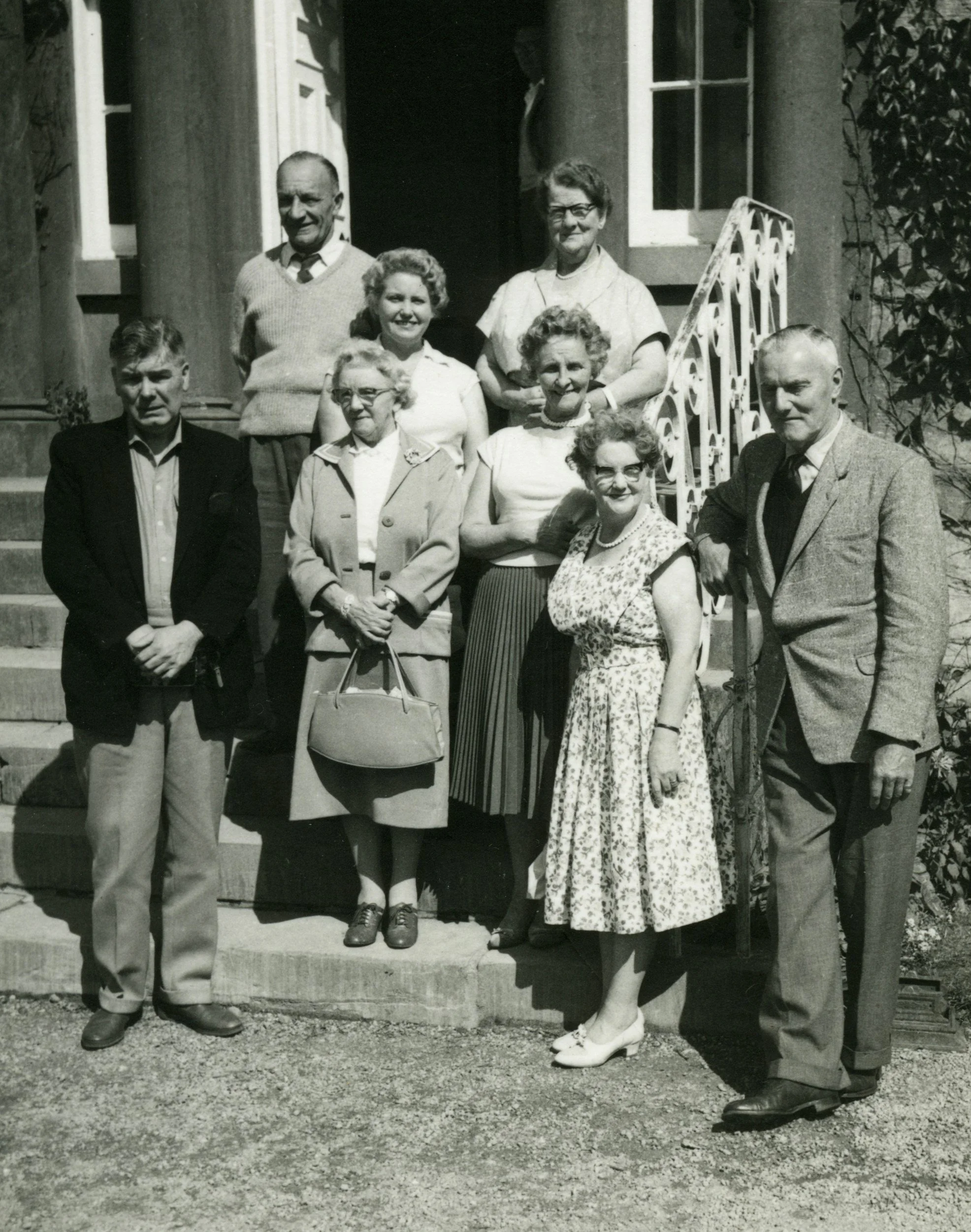 A black and white photograph of nine people, six women and three men, standing on the steps outside a house, dressed in vintage clothing from the mid-20th century.