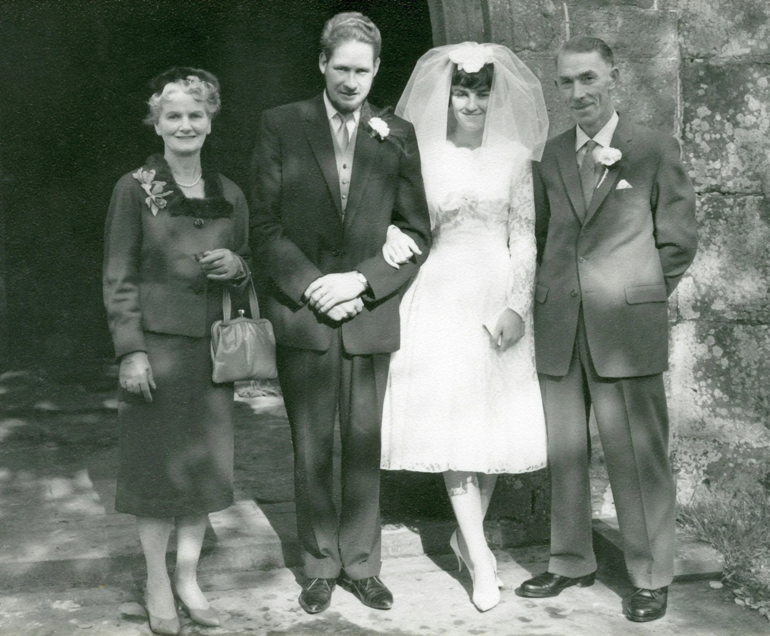 Black and white photo of a wedding scene with four people, including a bride in a white lace dress with a veil, and three others dressed in formal attire, standing outside against a stone wall.