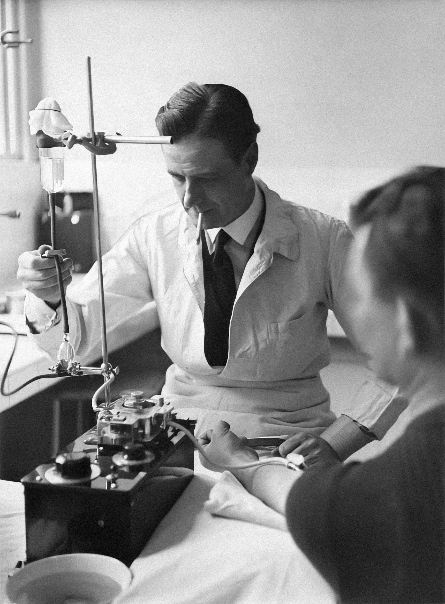 A black and white photograph of a man in a white lab coat conducting a scientific experiment, with a woman observing him. The man appears focused and is handling laboratory equipment, including a stand and some tubing.