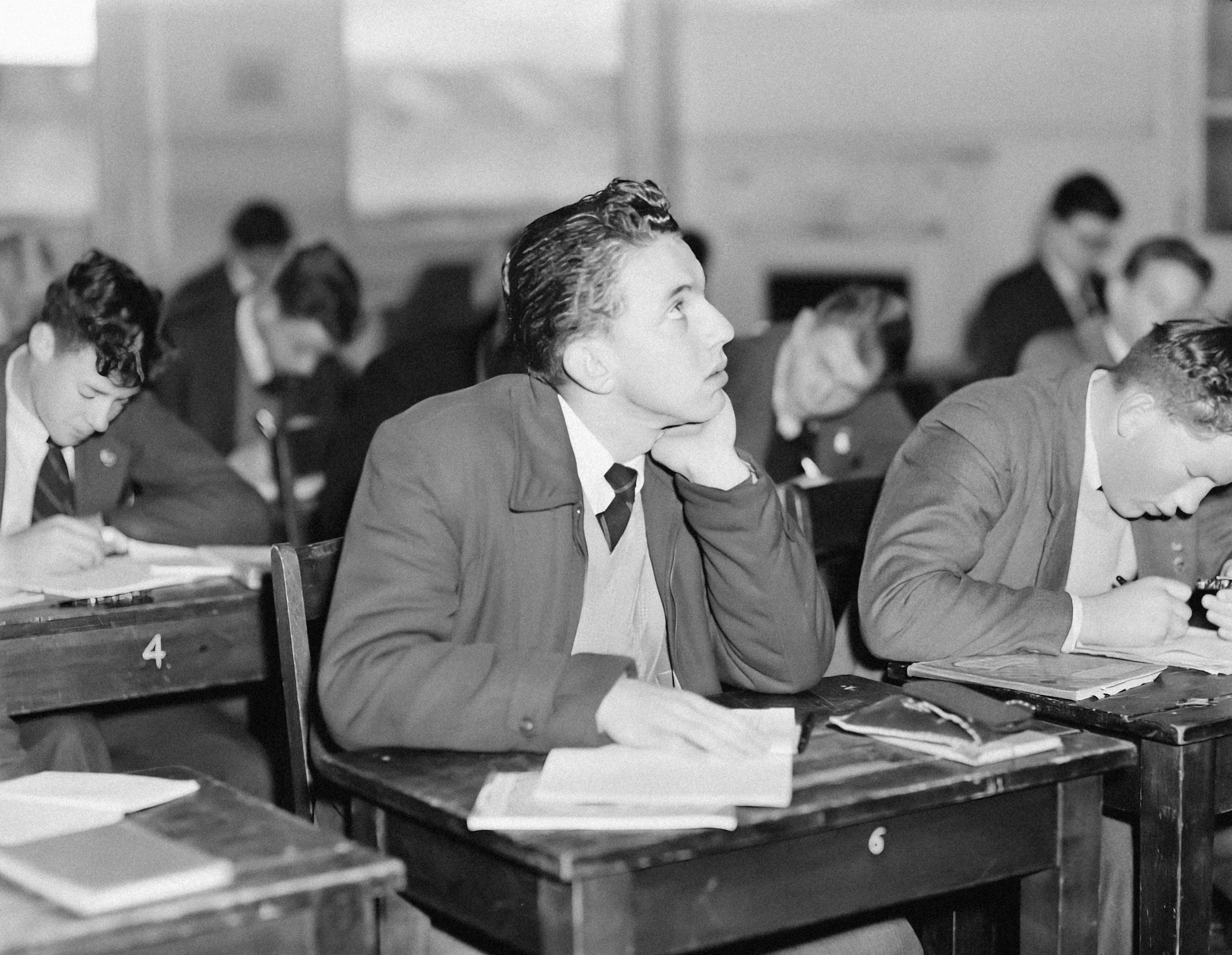 Students in a classroom, with a young man in focus resting his chin on his hand, looking up, surrounded by other students who are studying or writing.