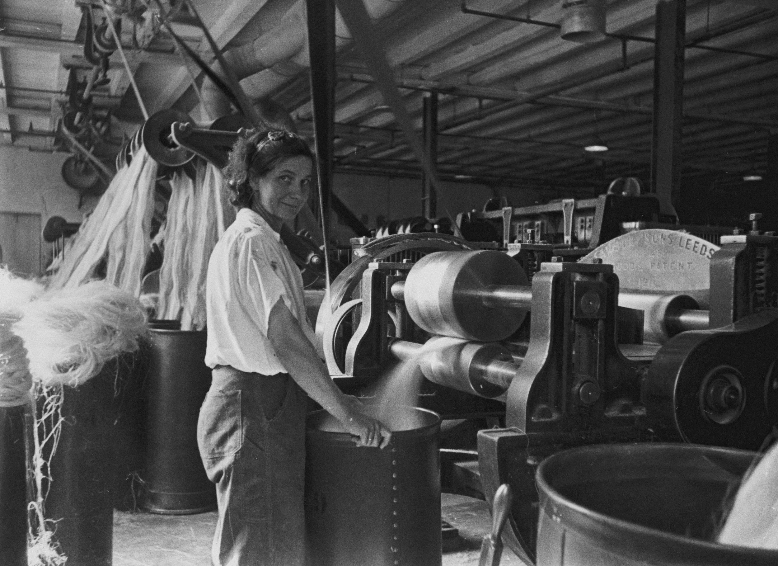 A woman working in a textile factory, operating a large industrial machine with spools of thread in the background.