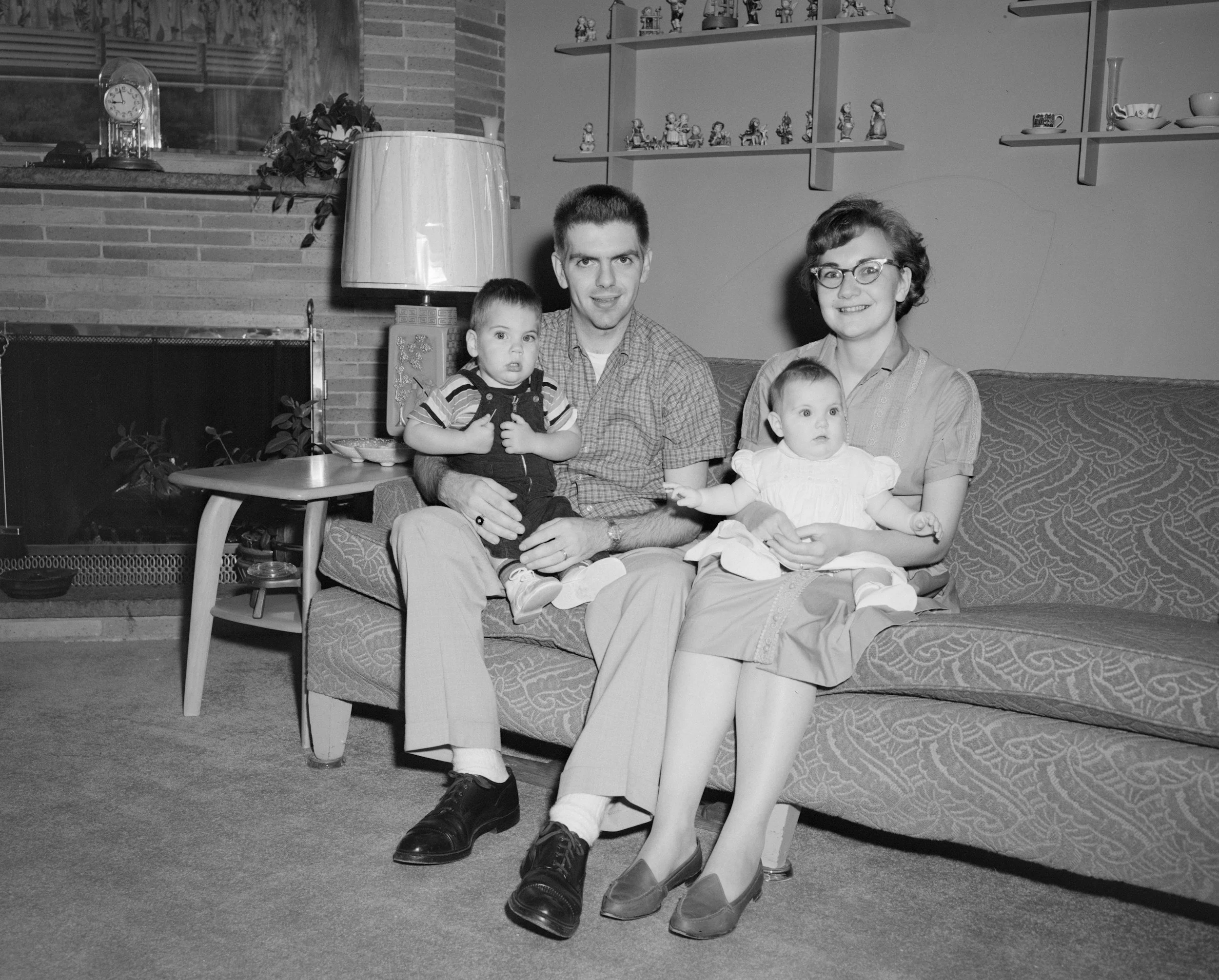 A black and white photo of a family of four sitting on a sofa in a living room. The father is on the left, holding a young boy, and the mother is on the right, holding a young girl. The room has a brick fireplace, shelves with small decorative items, and a window in the background.