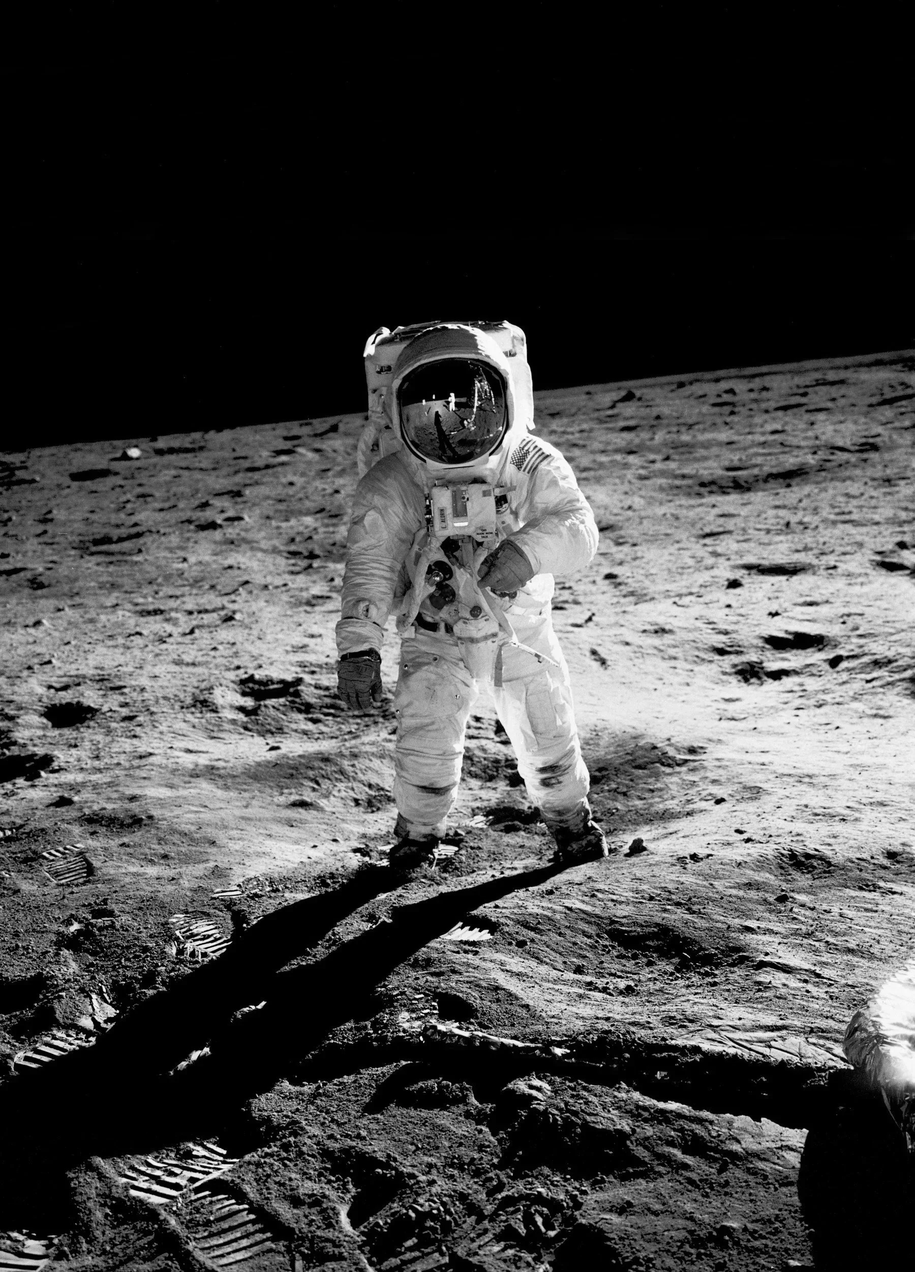 Black-and-white photo of an astronaut standing on the moon's surface, with footprints visible in the soil, and Earth reflected in the helmet visor.