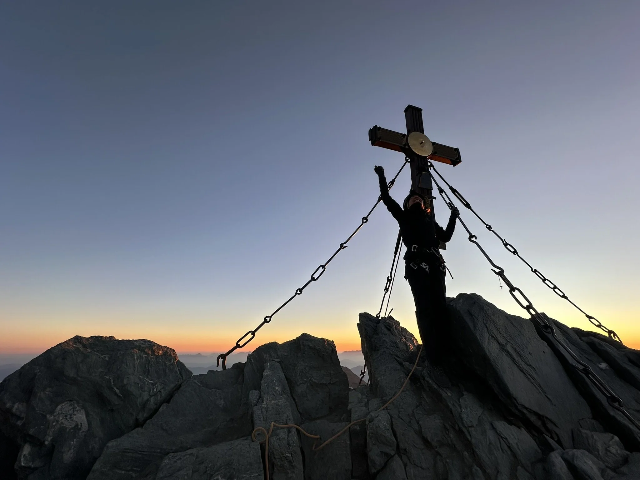 Person standing on rocky mountain peak at sunrise, holding onto a large wooden cross with chains.
