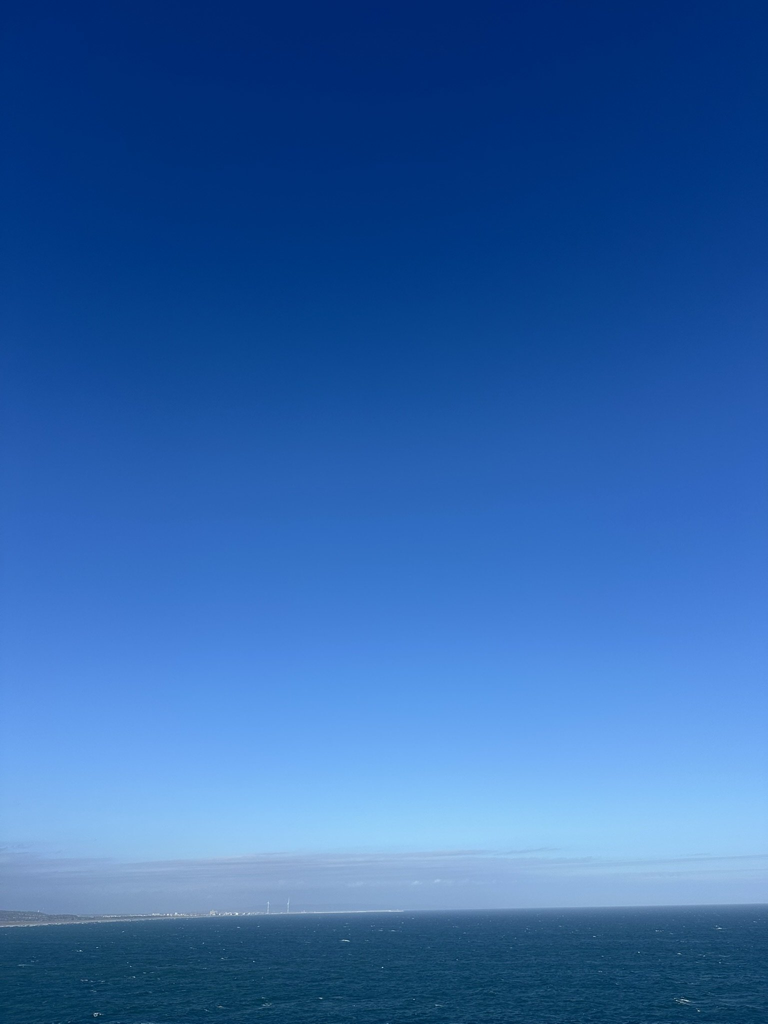 View of the ocean with a distant shoreline and wind turbines under a mostly clear blue sky.