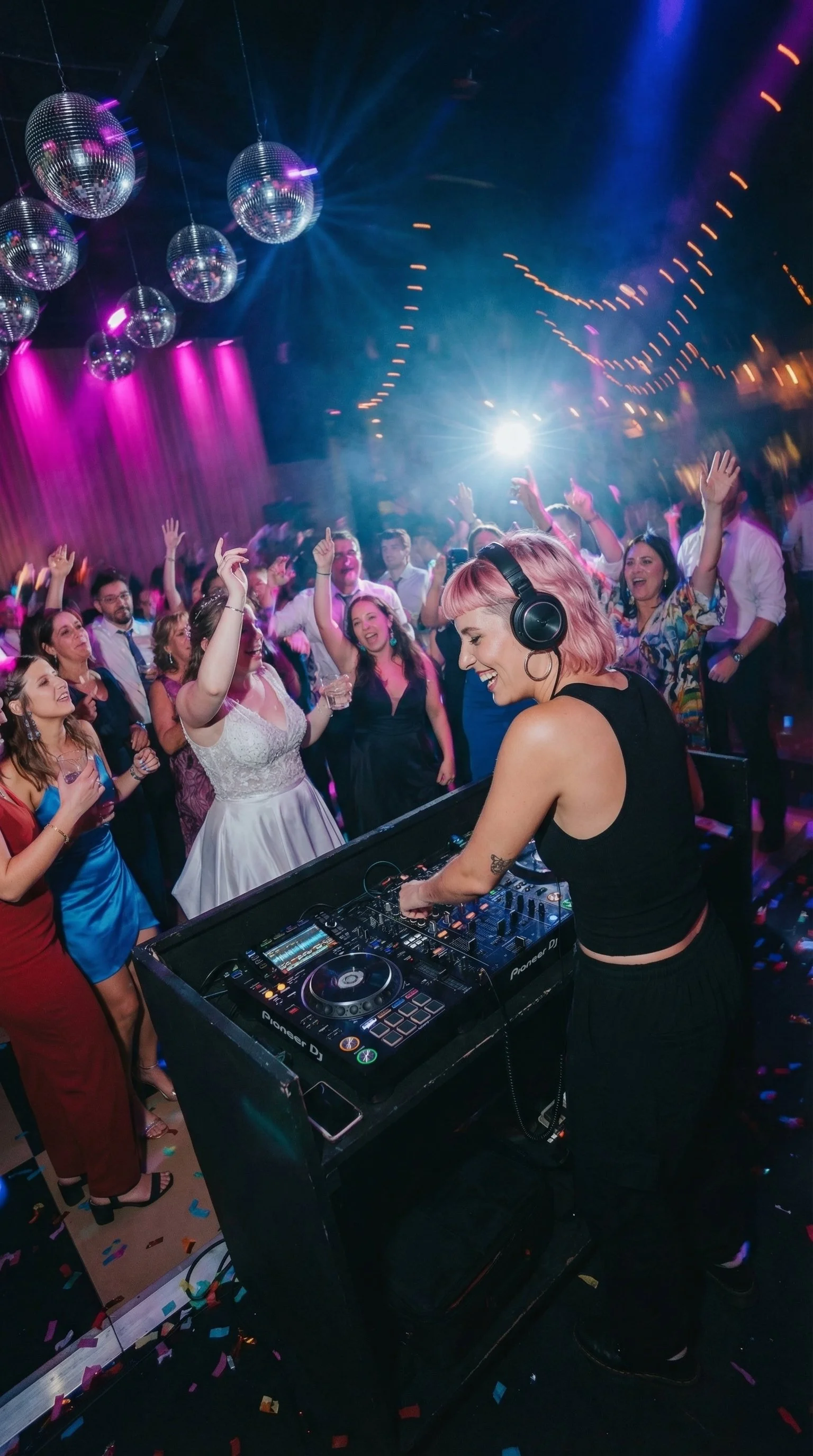 A female DJ with pink hair wearing headphones performs at a lively party with people dancing and celebrating under disco balls and colorful lights.
