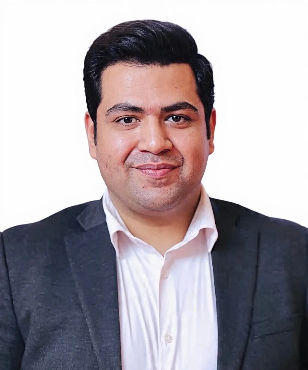 Portrait of a man with black hair wearing a white shirt and dark blazer, smiling at the camera with a plain white background.