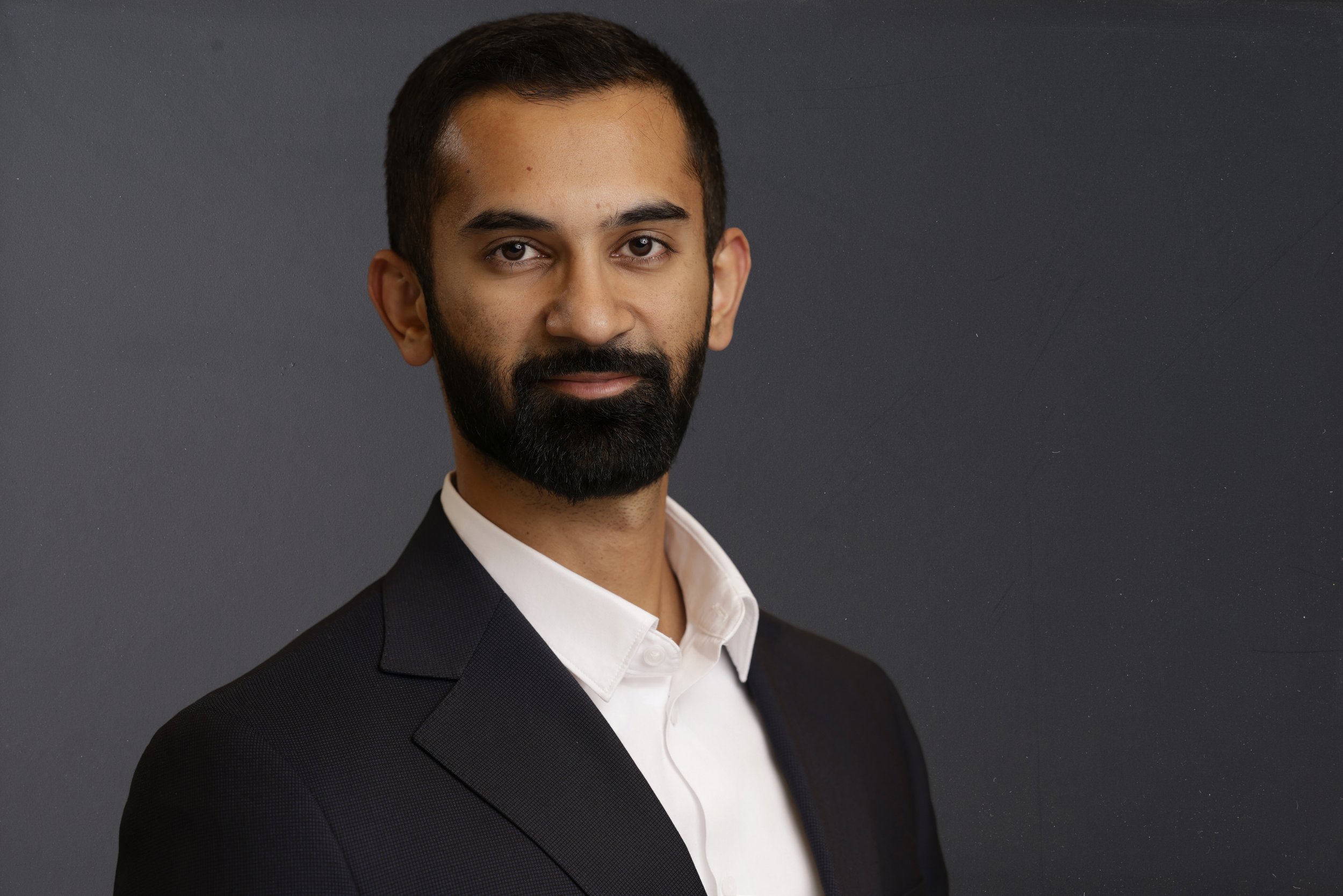 A professional man with a beard and short dark hair, wearing a black suit and white shirt, standing against a dark gray background.