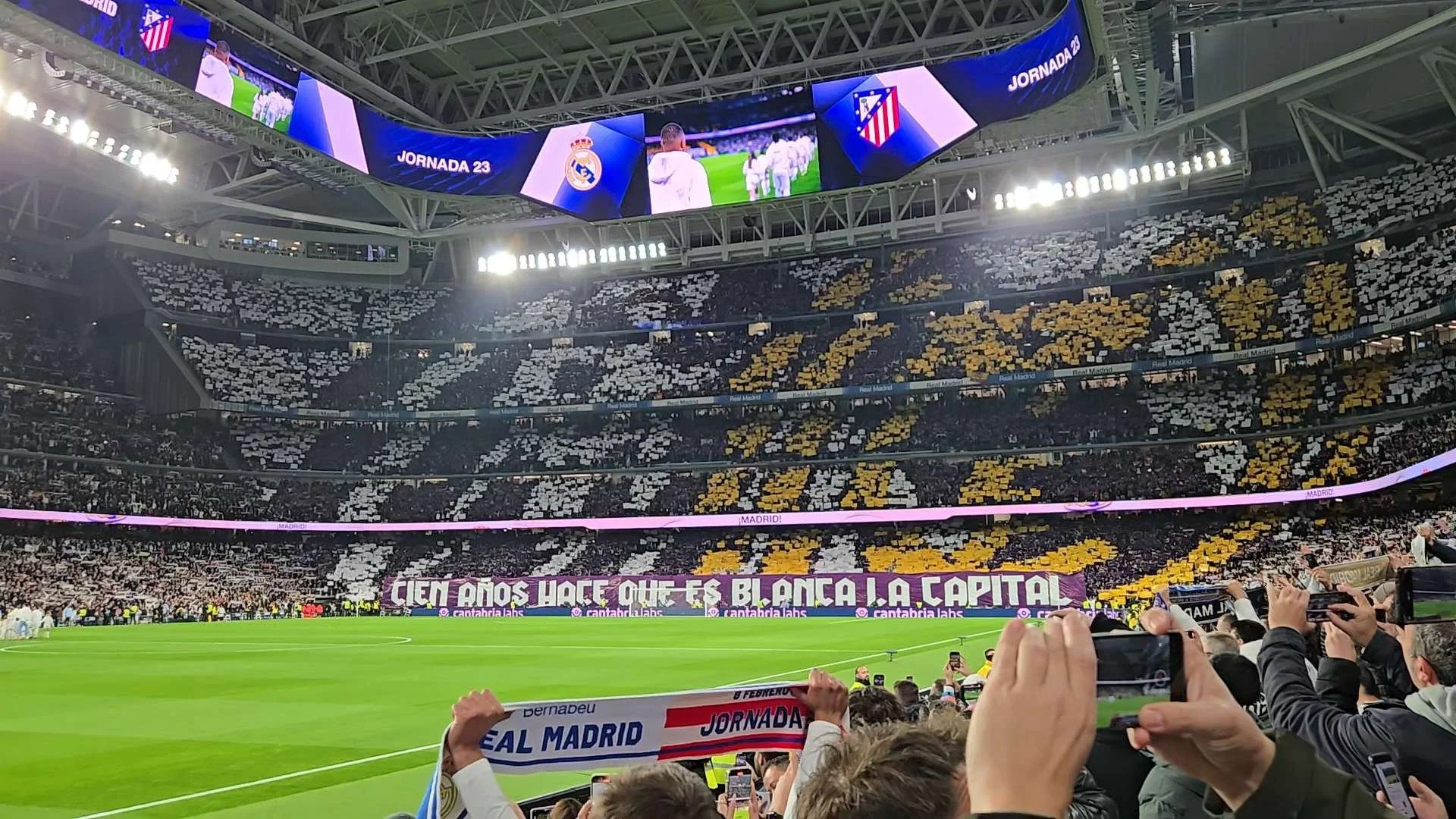 A soccer stadium filled with spectators, showing banners and crowd patterns in black, white, and yellow. The large banner in the stands reads 'CIEN AÑOS HACE QUE ES BLANCA! LA CAPITÁN.' The field is green with players and officials, and large digital