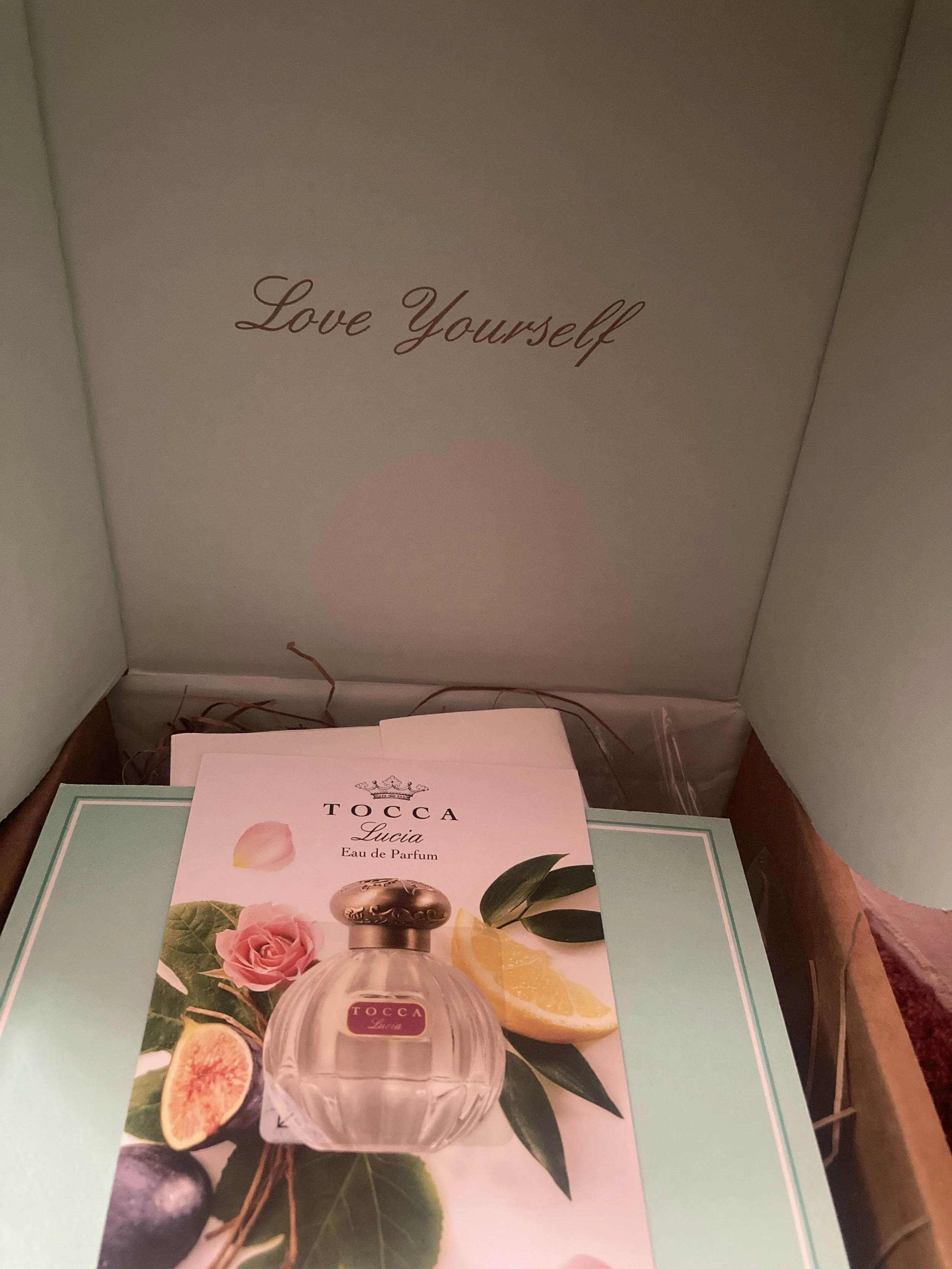 Open box with the phrase "Love Yourself" on the inside of the lid, containing a TOCCA Lucia perfume sample card with an illustration of the perfume bottle surrounded by figs, lemon, and leaves.