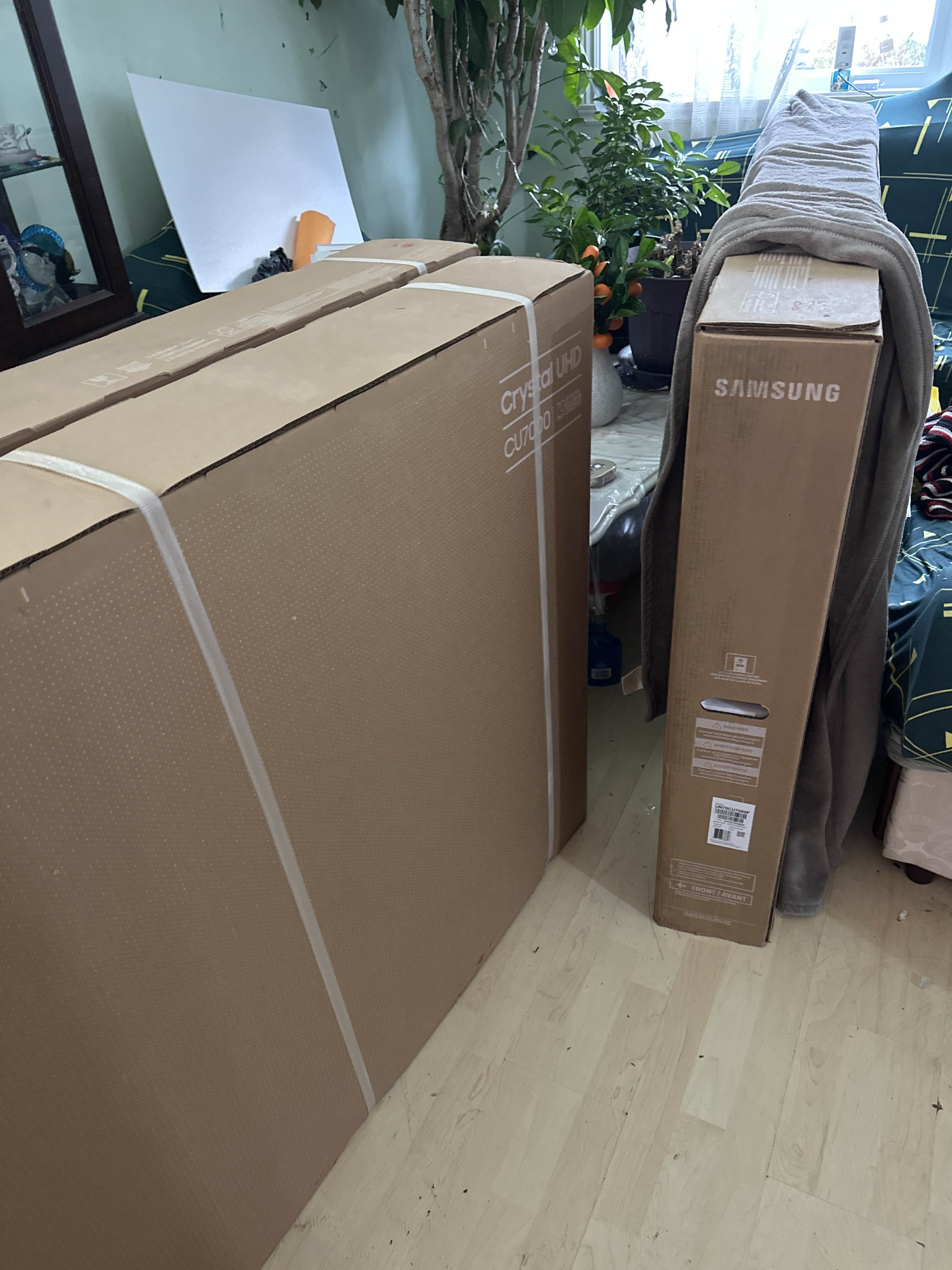 Two large boxed items, one labeled Samsung and the other Cryo HD, placed in a room with a wooden floor and near a plant, with some clutter and furniture visible in the background.