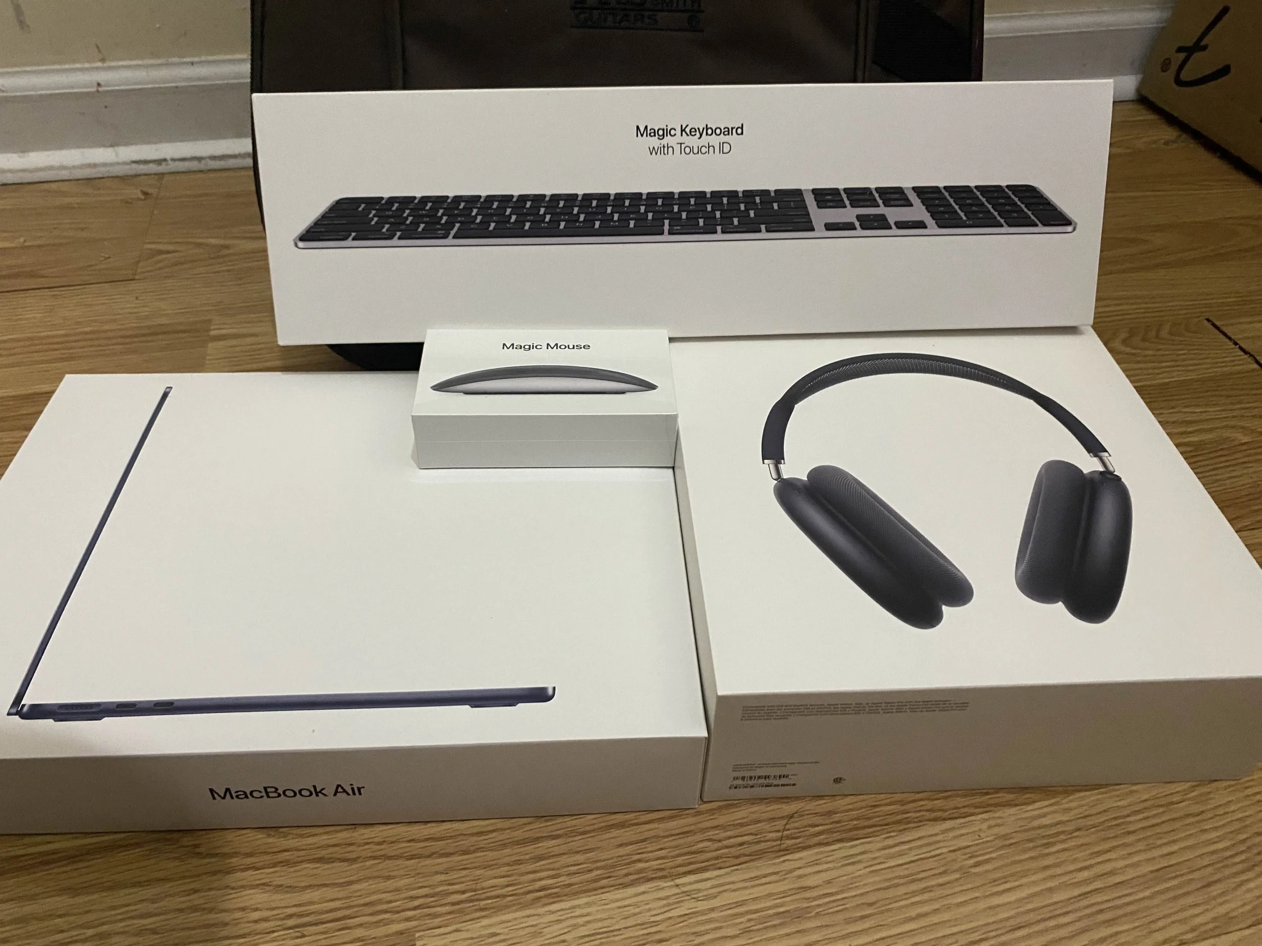 Unboxed Apple products including a MacBook Air, Magic Keyboard, Magic Mouse, and headphones on a wooden surface.