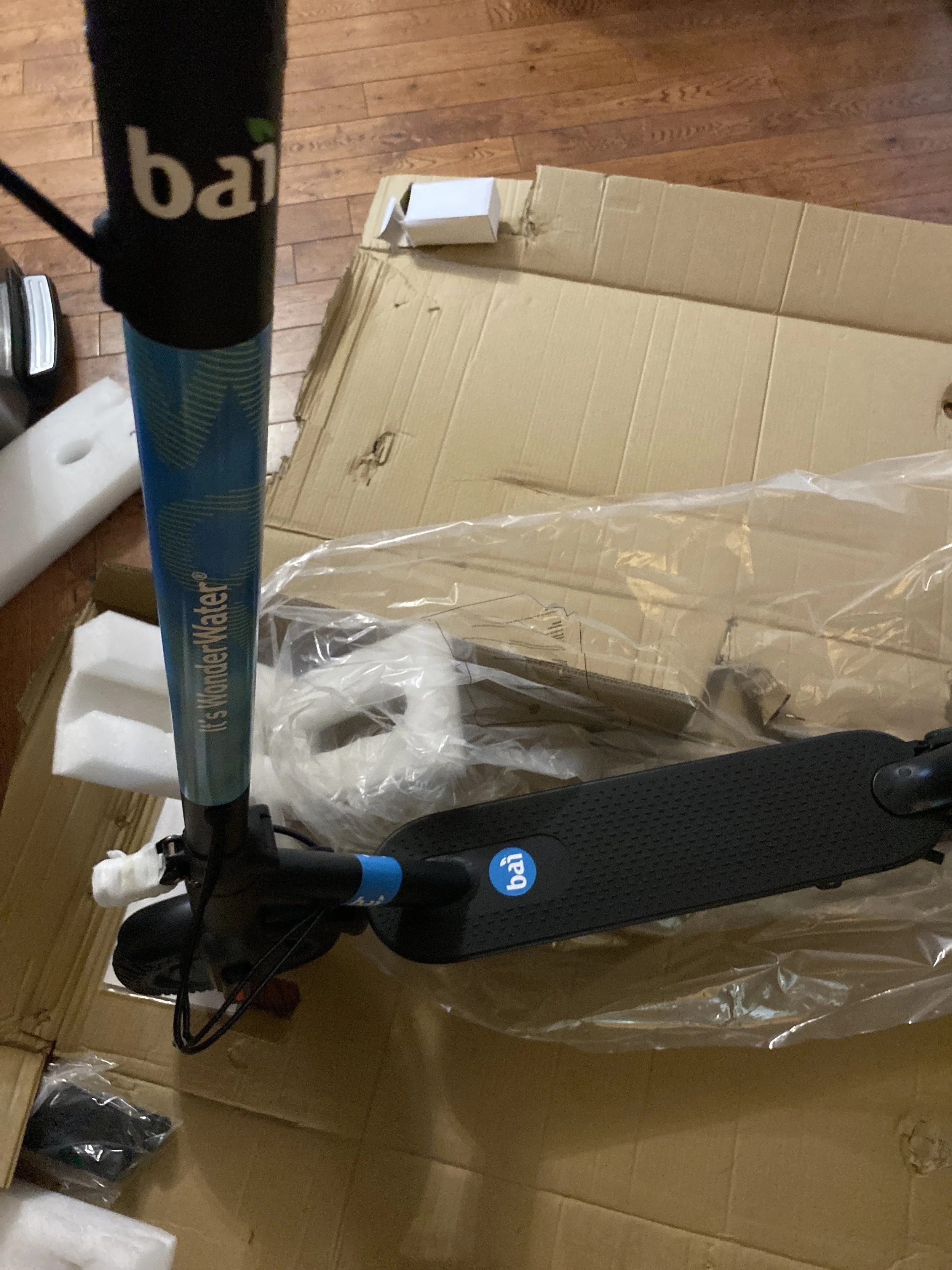 New black and blue electric scooter with white branding, partially unwrapped from plastic, on a wooden floor with packaging boxes and foam around.