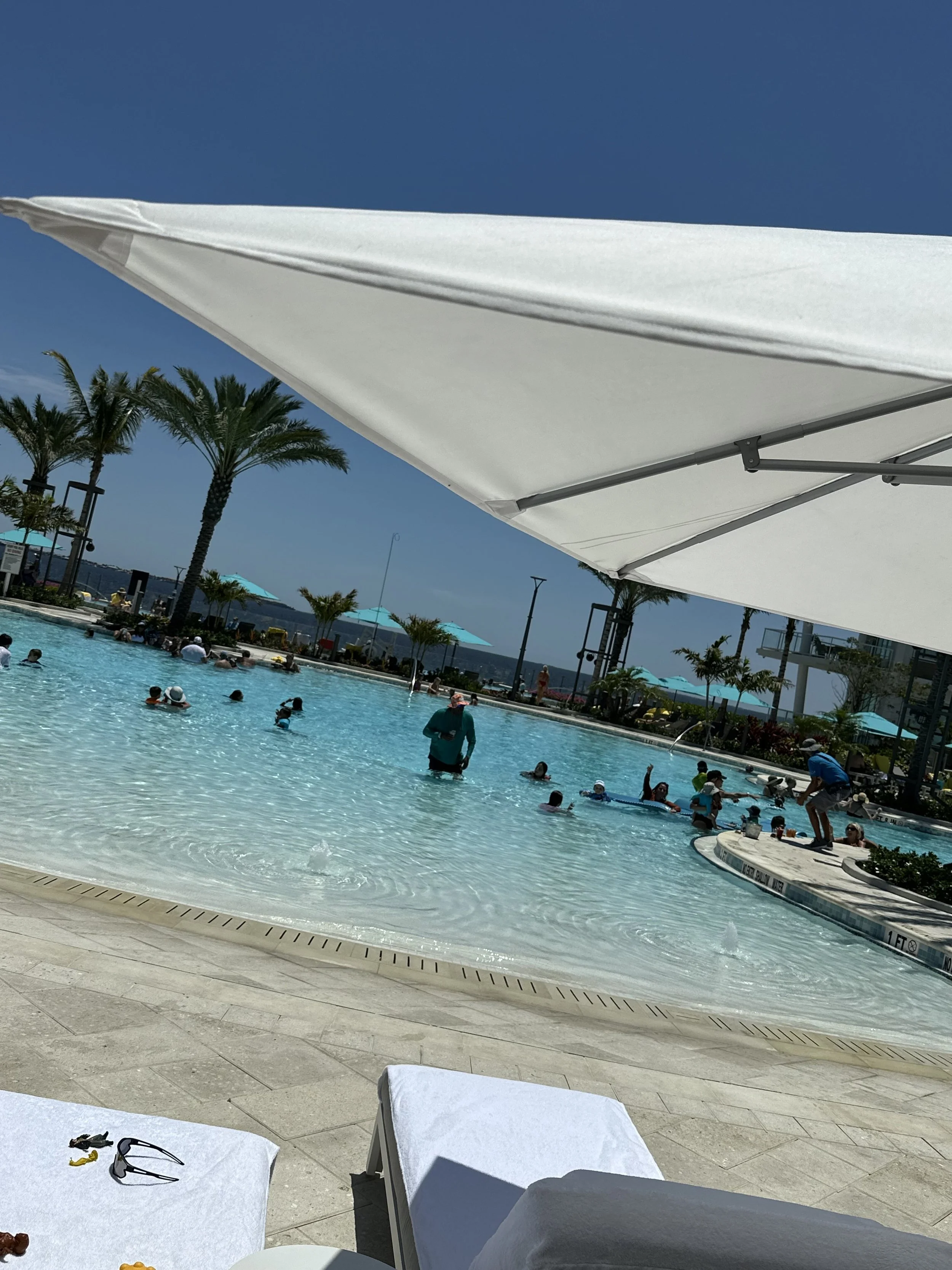 People enjoying a swimming pool under a clear blue sky, with large white umbrellas and palm trees in the background.