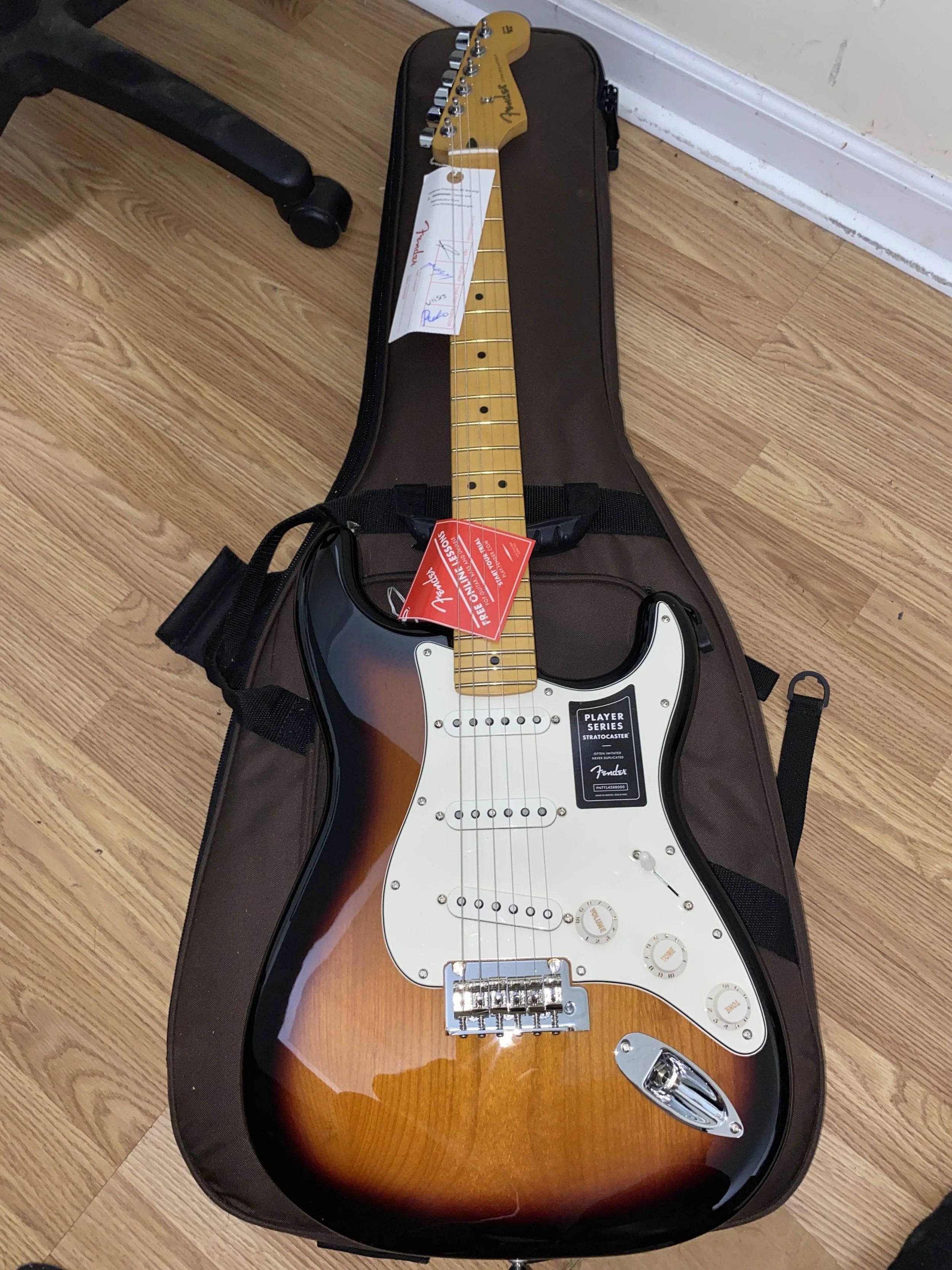 A Fender Stratocaster electric guitar with a sunburst finish resting on a soft gig bag on a wood floor, with tags and a name tag attached.