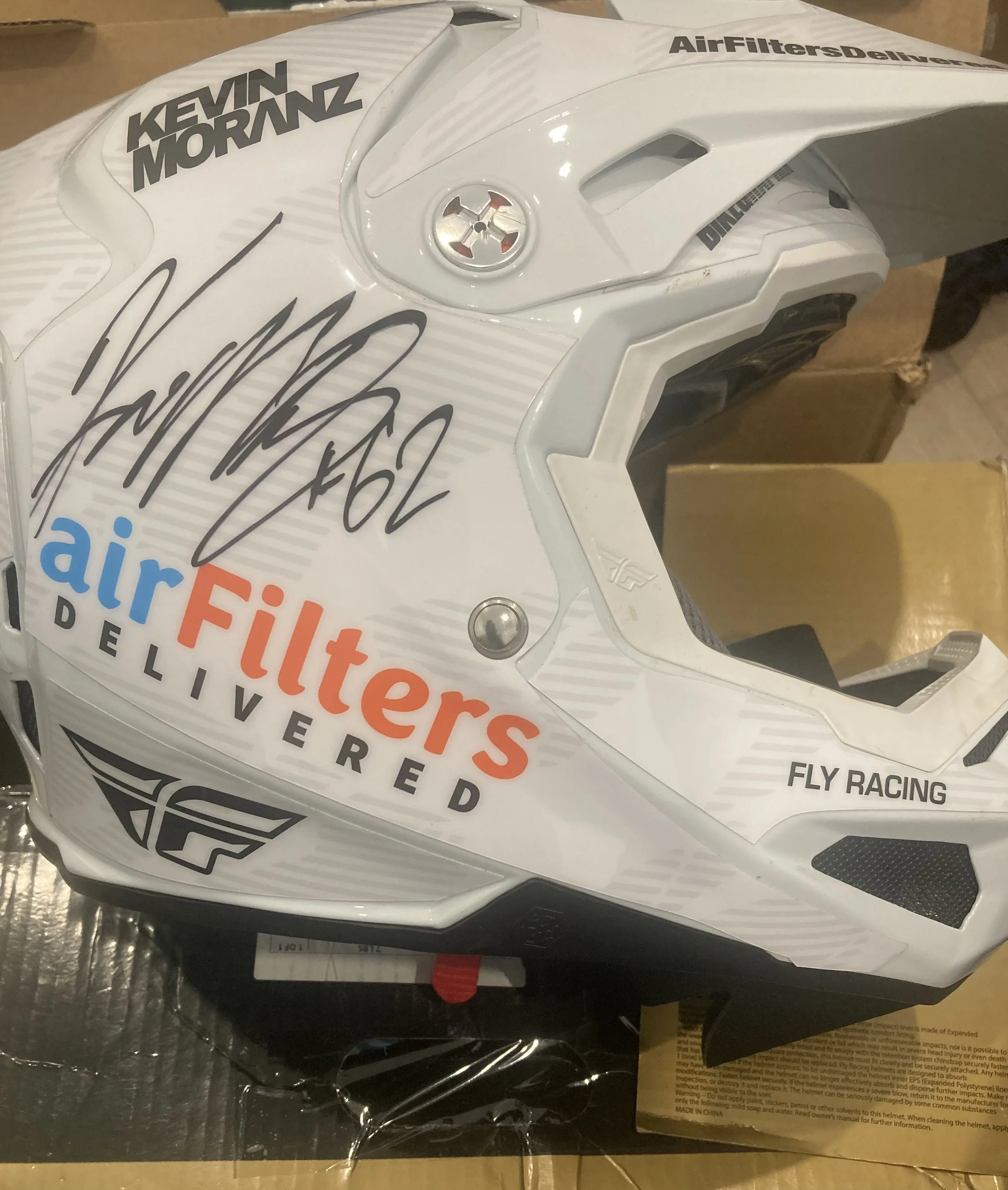 A white motocross helmet with signatures and logos, including Kevin Moranz and Air Filters Delivered, with black and red text and black graphics.
