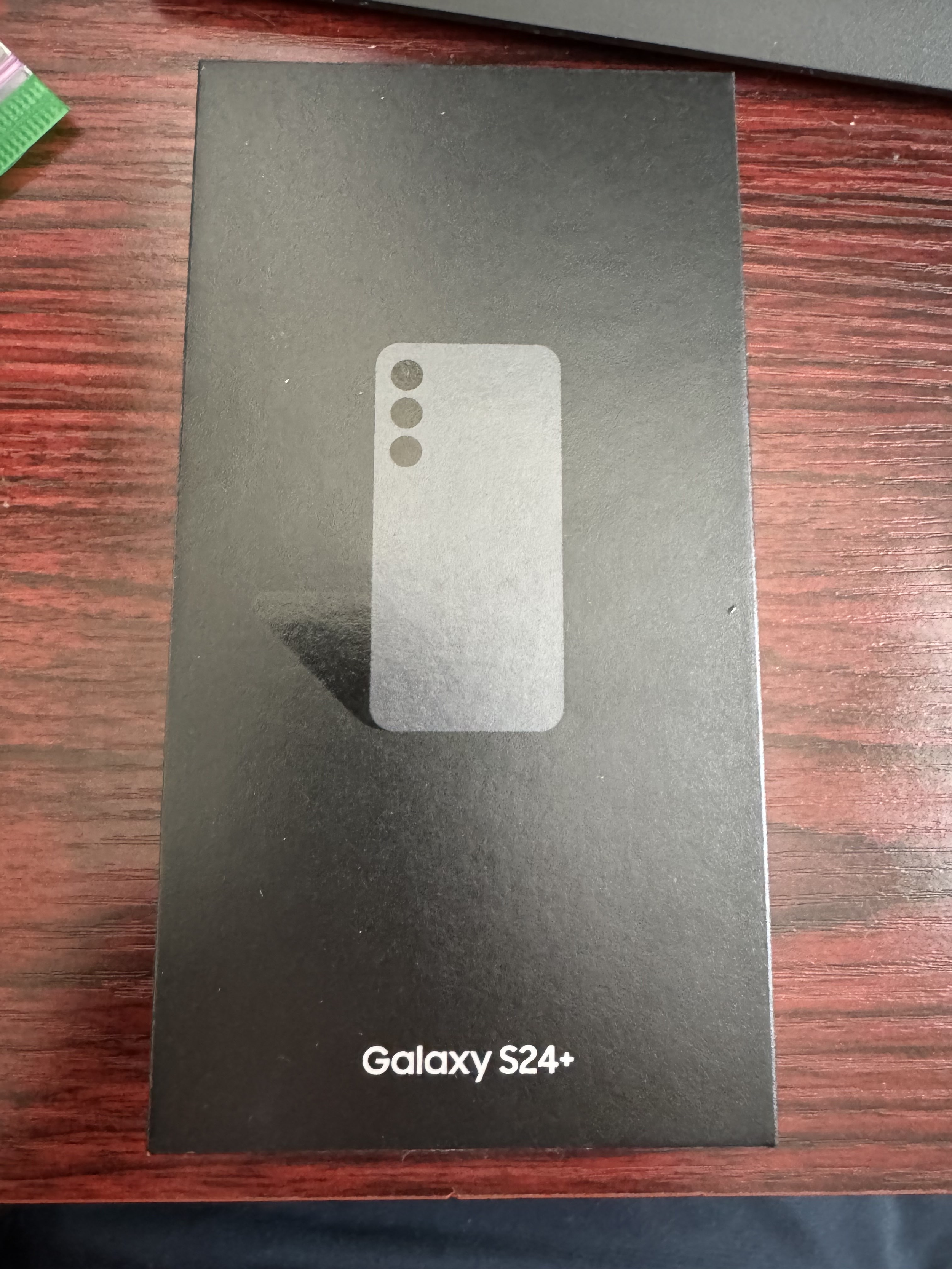 Black box packaging for Galaxy S24+ smartphone, with a simple graphic of the phone's silhouette and camera layout, placed on a wooden surface.