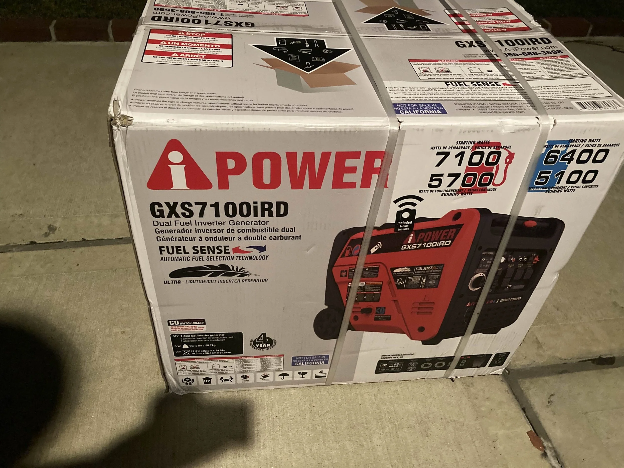 Boxed portable dual fuel inverter generator, model GXS7100iRD, with red and black color, placed on a concrete surface, secured with metal straps.