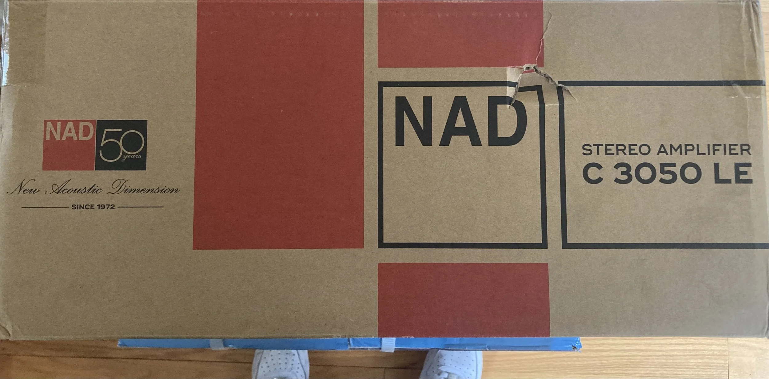 Brown cardboard box with black and red text and logos, indicating it contains NAD C 3050 LE stereo amplifier, celebrating NAD's 50 years since 1972, placed on a wooden floor.
